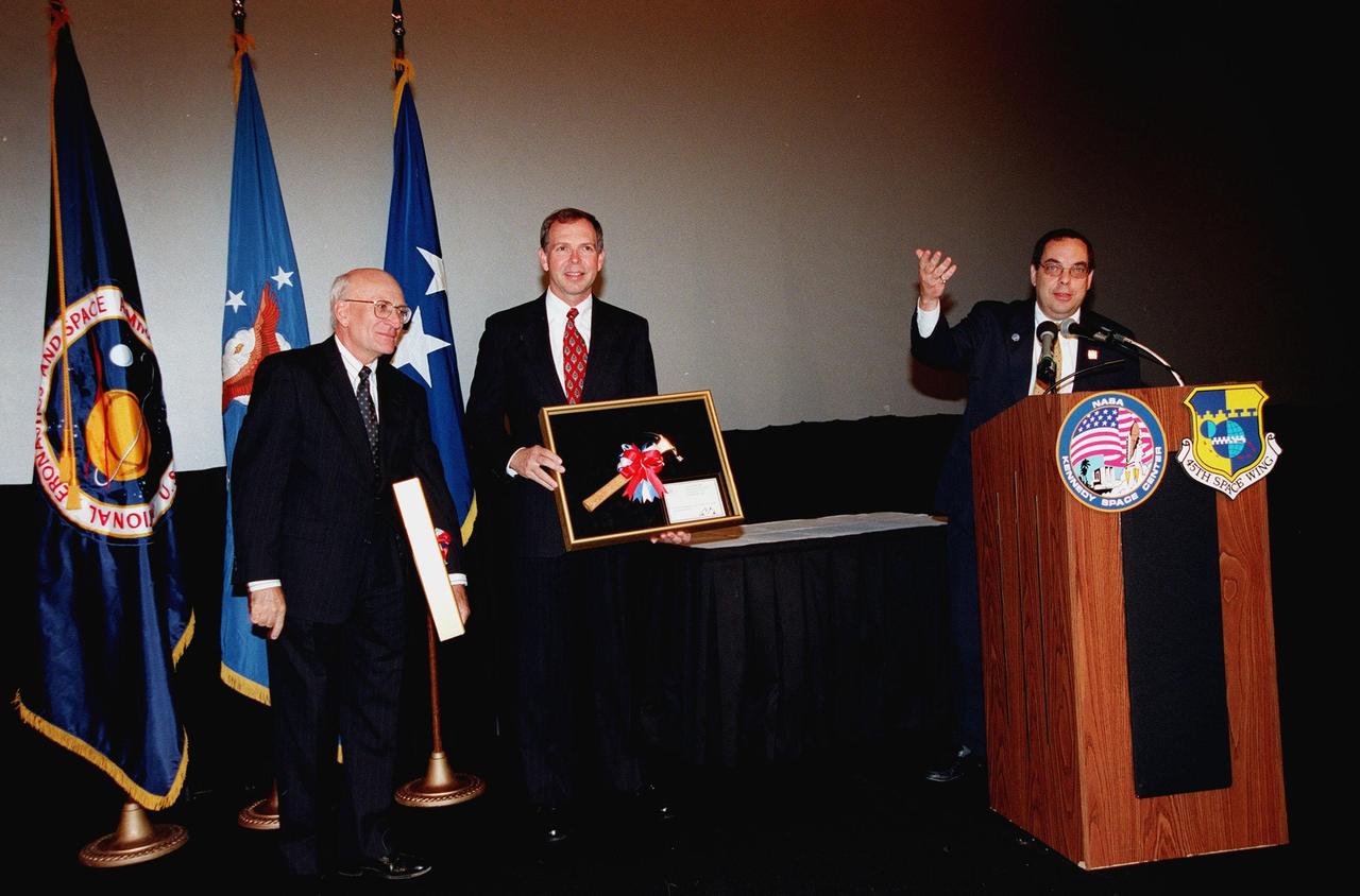 Ed Gormel (left) and Chris Fairey (center) display the Hammer Award they received at a special presentation in the IMAX 2 Theater in the Kennedy Space Center Visitor Complex. The Hammer Award is Vice President Al Gore's special recognition of teams of federal employees who have made significant contributions in support of the principles of the National Partnership for Reinventing Government. At the podium is Morley Winograd director of the National Partnership for Reinventing Government, who presented the award. This Hammer Award acknowledges the accomplishments of a joint NASA and Air Force team that established the Joint Base Operations and Support Contract (J-BOSC) Source Evaluation Board (SEB). Fairey and Gormel are co-chairs of the SEB. The team developed and implemented the acquisition strategy for establishing a single set of base operations and support service requirements for KSC, Cape Canaveral Air Station and Patrick Air Force Base