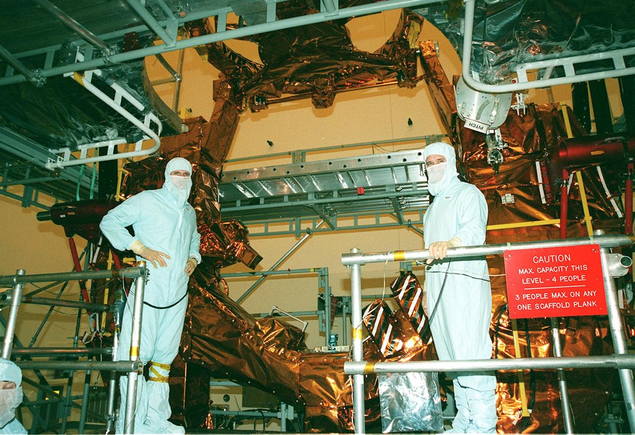 Standing on a workstand in the Payload Hazardous Servicing Facility, STS-103 Mission Specialists Steven L. Smith and John M. Grunsfield (Ph.D.) pose for the camera while standing in front of the base of the Flight Support System, to be used for repair of the Hubble Space Telescope, the primary mission on STS-103. The crew are at KSC to take part in a Crew Equipment Interface Test. Other members of the crew are Commander Curtis L. Brown Jr., Pilot Scott J. Kelly, and Mission Specialists C. Michael Foale (Ph.D.), Claude Nicollier of Switzerland, and Jean-François Clervoy of France. Nicollier and Clervoy are with the European Space Agency. Mission STS-103 is a "call-up" due to the need to replace portions of the pointing system, the gyros, which have begun to fail on the Hubble Space Telescope. Although Hubble is operating normally and conducting its scientific observations, only three of its six gyroscopes are working properly. The gyroscopes allow the telescope to point at stars, galaxies and planets. The STS-103 crew will not only replace gyroscopes, it will also replace a Fine Guidance Sensor and an older computer with a new enhanced model, an older data tape recorder with a solid-state digital recorder, a failed spare transmitter with a new one, and degraded insulation on the telescope with new thermal insulation. The crew will also install a Battery Voltage/Temperature Improvement Kit to protect the spacecraft batteries from overcharging and overheating when the telescope goes into a safe mode. The scheduled launch date in October is under review