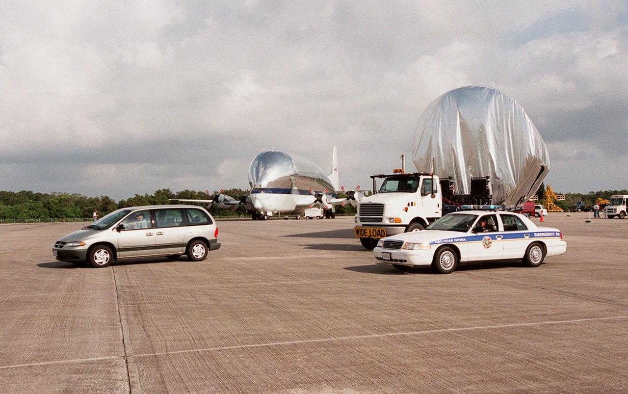 KENNEDY SPACE CENTER, FLA. -- Escort vehicles prepare to leave the Shuttle Landing Facility with the S1 truss (at right) on its trek to the Operations and Checkout Building. Manufactured by the Boeing Co. in Huntington Beach, Calif., this component of the ISS is the first starboard (right-side) truss segment, whose main job is providing structural support for the orbiting research facility's radiator panels that cool the Space Station's complex power system. The S1 truss segment also will house communications systems, external experiment positions and other subsystems. Primarily constructed of aluminum, the truss segment is 45 feet long, 15 feet wide and 6 feet tall. When fully outfitted, it will weigh 31,137 pounds. The truss is slated for flight in 2001. The truss arrived at KSC aboard NASA's Super Guppy, seen in the background. The aircraft is uniquely built with a 25-foot diameter fuselage designed to handle oversized loads and a "fold-away" nose that opens 110 degrees for cargo loading. A system of rails in the cargo compartment, used with either Guppy pallets or fixtures designed for specific cargo, makes cargo loading simple and efficient. Rollers mounted in the rails allow pallets or fixtures to be moved by an electric winch mounted beneath the cargo floor. Automatic hydraulic lock pins in each rail secure the pallet for flight