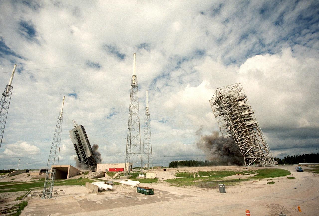 KENNEDY SPACE CENTER, FLA. — Two 34-year-old towers on Launch Complex 41, Cape Canaveral Air Station, fall to the ground amid the black smoke from explosives set to topple them. Weighing two million pounds, the umbilical tower (left) was approximately 200 feet high. The taller 300-foot Mobile Service Tower (right) weighs five million pounds. About 200 pounds of linear-shaped charges were used to topple the towers so that the materials can be recycled. Adjacent to the towers are lightning protection structures, which will remain on the site. The towers are being demolished to make room for Lockheed Martin's 14-acre Vehicle Integration Facility (VIF), under construction. The implosion and removal of the tower debris is expected to be completed in two months. The VIF will be used for Lockheed Martin's Atlas V Launch System.