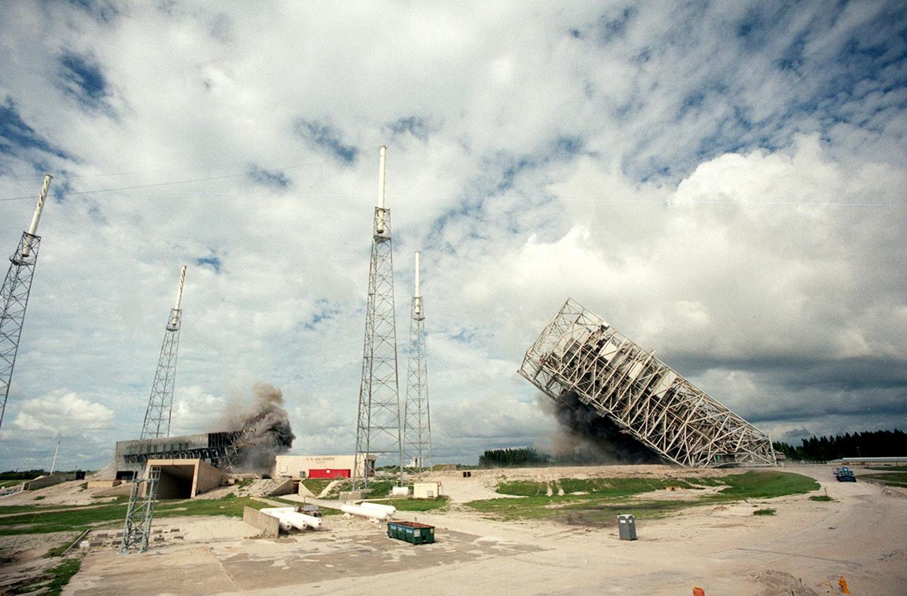 KENNEDY SPACE CENTER, FLA. — Two 34-year-old towers on Launch Complex 41, Cape Canaveral Air Station, fall to the ground amid the black smoke from explosives set to topple them. Weighing two million pounds, the umbilical tower (left) was approximately 200 feet high. The taller 300-foot Mobile Service Tower (right), still falling, weighs five million pounds. About 200 pounds of linear-shaped charges were used to topple the towers so that the materials can be recycled. Adjacent to the towers are lightning protection structures, which will remain on the site. The towers are being demolished to make room for Lockheed Martin's 14-acre Vehicle Integration Facility (VIF), under construction. The implosion and removal of the tower debris is expected to be completed in two months. The VIF will be used for Lockheed Martin's Atlas V Launch System.