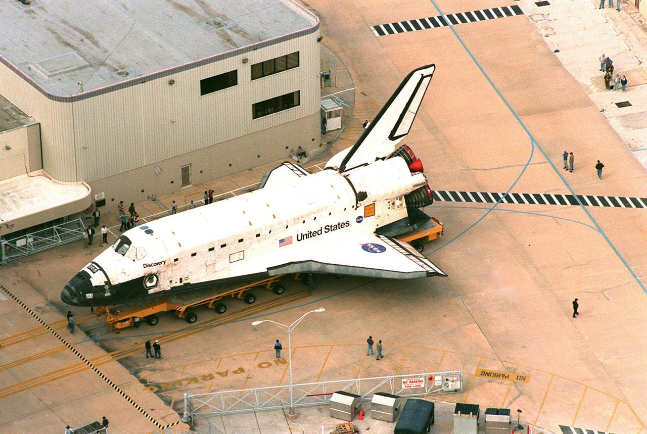 In this aerial view, the orbiter Discovery is out of the Orbiter Processing Facility (OPF) bay 1 and rolling back before onto the tow-way for its rollover to the Vehicle Assembly Building where it will be mated with an external tank and solid rocket boosters before its launch on mission STS-103. The launch date is currently under review for early December. STS-103, the third Hubble Space Telescope servicing mission, is a "call-up" due to the need to replace portions of the pointing system, the gyros, which have begun to fail on the Hubble Space Telescope. Although Hubble is operating normally and conducting its scientific observations, only three of its six gyroscopes are working properly. The gyroscopes allow the telescope to point at stars, galaxies and planets. The STS-103 crew will also be replacing a Fine Guidance Sensor and an older computer with a new enhanced model, an older data tape recorder with a solid-state digital recorder, a failed spare transmitter with a new one, and degraded insulation on the telescope with new thermal insulation. The crew will also install a Battery Voltage/Temperature Improvement Kit to protect the spacecraft batteries from overcharging and overheating when the telescope goes into a safe mode