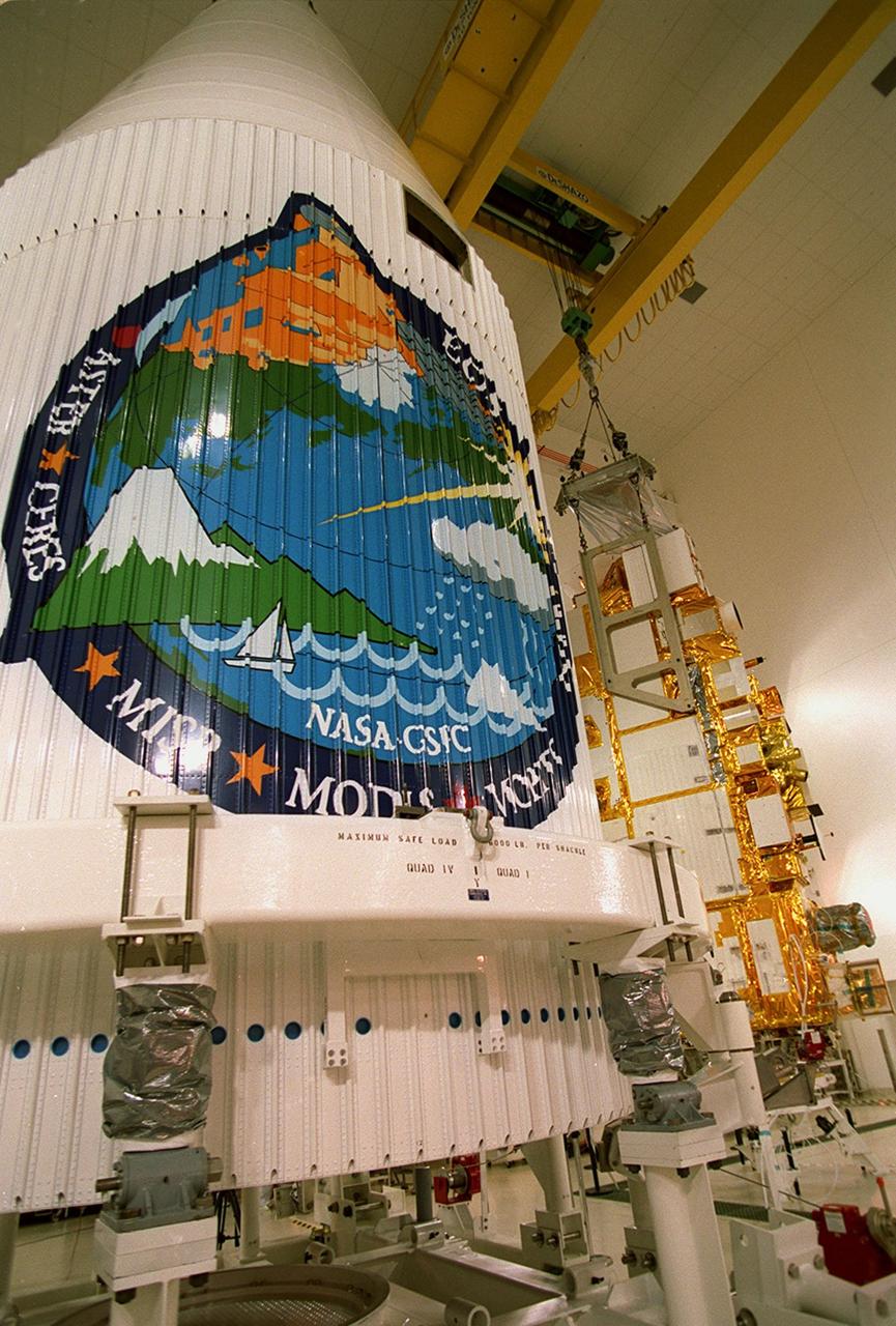 The rocket faring (left) displays a logo of the many science instruments that make up NASA's Terra spacecraft (background). When fully assembled, Terra will be encapsulated in the faring before launch, scheduled for Dec. 16 aboard a Lockheed Martin Atlas IIAS rocket from Space Launch Complex 3 East at Vandenberg AFB, Calif. Terra comprises five state-of-the-art sets of instruments that will collect data for continuous, long-term records of the state of Earth's land, oceans and atmosphere. Together with data from other satellite systems launched by NASA and other countries, it will inaugurate a new self-consistent data record that will be gathered over the next 15 years. From an altitude of 438 miles, Terra will circle the Earth 16 times a day from pole to pole (98 degree inclination), crossing the equator at 10:30 a.m. The five Terra instruments will operate by measuring sunlight reflected by the Earth and heat emitted by the Earth