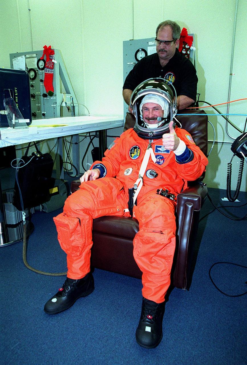 In the Operations and Checkout Building, STS-103 Commander Curtis L. Brown Jr. signals he's ready for launch while having his launch and entry suit checked by a suit technician. Other crew members are Pilot Scott J. Kelly and Mission Specialists Steven L. Smith, C. Michel Foale (Ph.D.), John M. Grunsfeld (Ph.D.), Claude Nicollier of Switzerland and Jean-François Clervoy of France. Nicollier and Clervoy are with the European Space Agency. The STS-103 mission, to service the Hubble Space Telescope, is scheduled for launch Dec. 17 at 8:47 p.m. EST from Launch Pad 39B. Mission objectives include replacing gyroscopes and an old computer, installing another solid state recorder, and replacing damaged insulation in the telescope. After the 8-day, 21-hour mission, Discovery is expected to land at KSC Sunday, Dec. 26, at about 6:30 p.m. EST