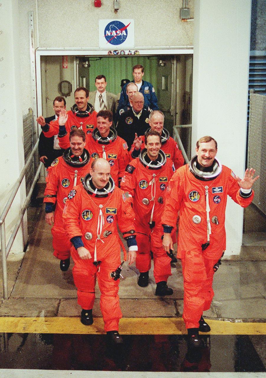 The STS-103 crew smile and wave to onlookers as they walk out of the Operations and Checkout Building enroute to Launch Pad 39B and liftoff of Space Shuttle Discovery. In their orange launch and entry suits, they are (front row) Pilot Scott J. Kelly and Commander Curtis L. Brown Jr., (second row) Mission Specialists John M. Grunsfeld (Ph.D.) and Jean-Francois Clervoy of France, (third row) C. Michael Foale (Ph. D.) and Claude Nicollier of Switzerland, and bringing up the rear, Steven L. Smith. The STS-103 mission, to service the Hubble Space Telescope, is scheduled for launch Dec. 17 at 8:47 p.m. EST from Launch Pad 39B. Mission objectives include replacing gyroscopes and an old computer, installing another solid state recorder, and replacing damaged insulation in the telescope. After the 8-day, 21-hour mission, Discovery is expected to land at KSC Sunday, Dec. 26, at about 6:30 p.m. EST