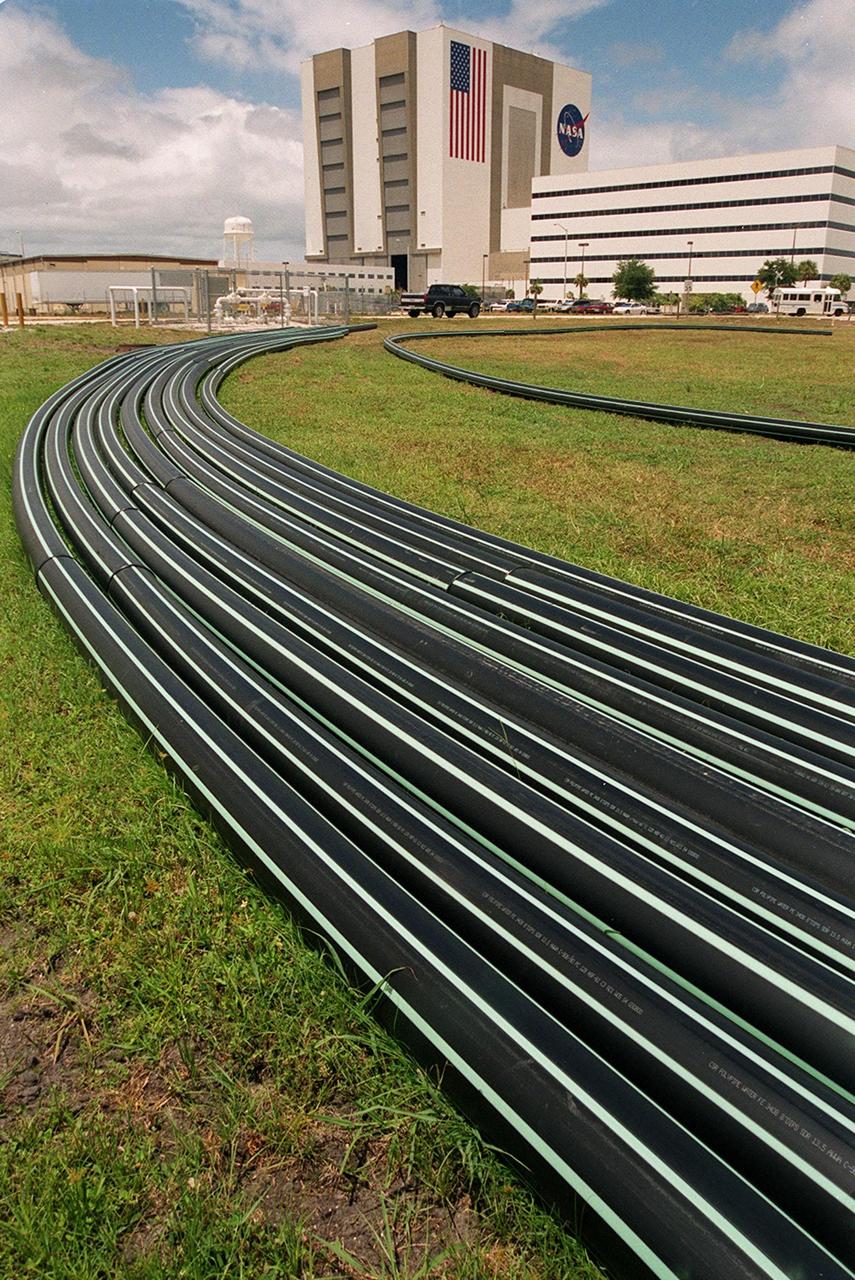 A bundle of flexible pipes arcing toward the Vehicle Assembly Building (left) and Operations Support Building (right) presents an artistic design to travelers on nearby Kennedy Parkway and Saturn Causeway