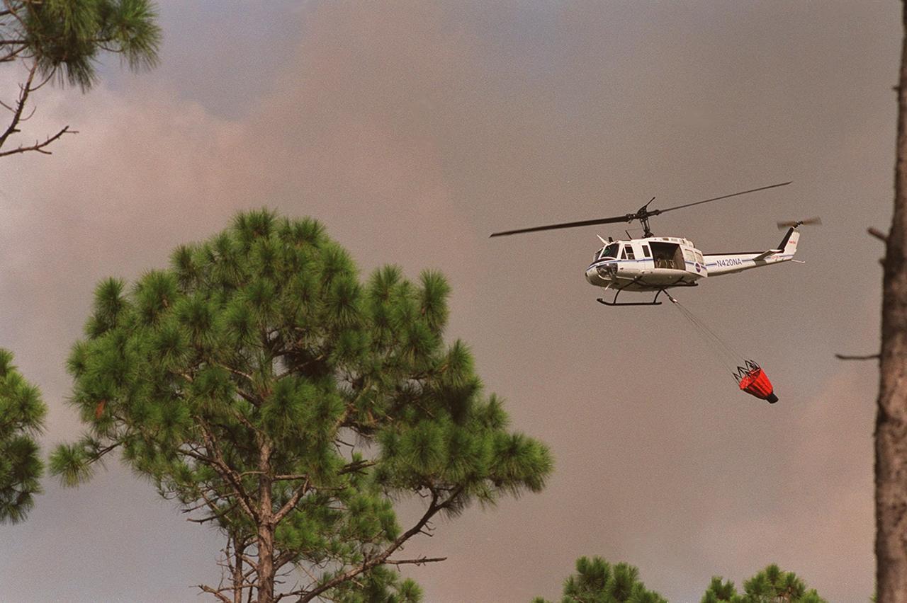 A NASA helicopter flies toward the source of the smoke to drop its load of water on a wildfire at KSC. Before being extinguished, the fire burned about 20 acres at a site near gate 2C on Kennedy Parkway North (route 3)