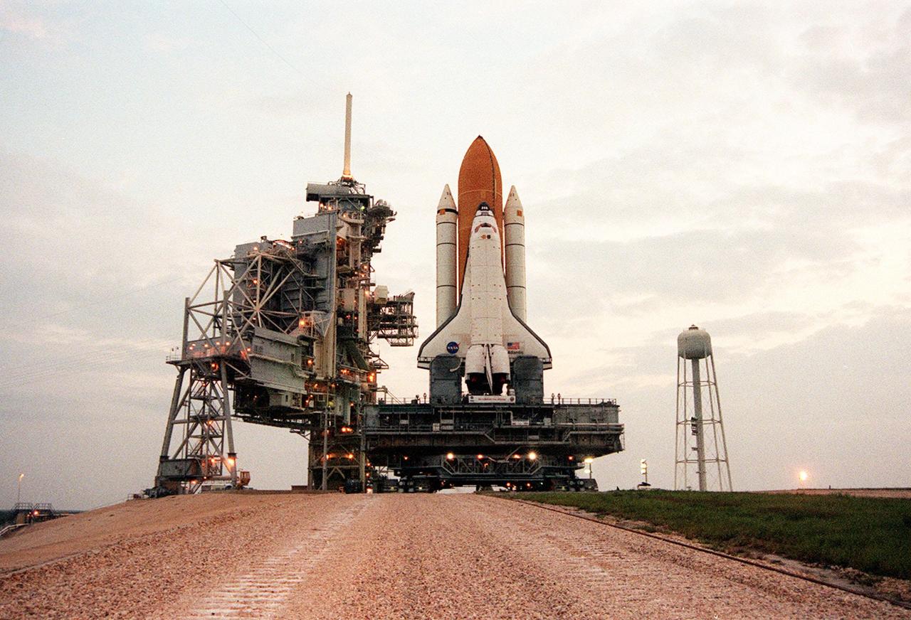 KENNEDY SPACE CENTER, Fla. --  The sun creeps over the horizon (right) as Space Shuttle Atlantis, atop the Mobile Launcher Platform and Crawler-Transporter, arrives at Launch Pad 39B for preparations before launch. It started its 8-hour rollout from the Vehicle Assembly Building high bay 2 (Safe Haven) at 11:20 p.m., Aug. 13. At left is the Rotating Service Structure in its open position. Towering above the RSS (and attached to the Fixed Service Structure) is the 80-foot tall lightning mast. At right is the 290-foot high water tank that holds 300,000 gallons of water. It is part of the sound suppression system at the pad. Atlantis is scheduled for launch Sept. 8 at 8:31 a.m. EDT on mission STS-106.