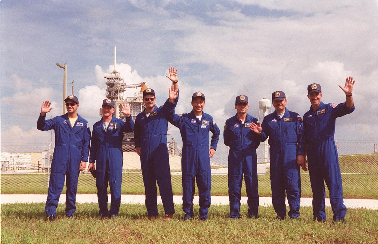 While meeting with family on the day before launch, the STS-106 crew poses for a photo. Waving, left to right, are Mission Specialist Richard A. Mastracchio, Commander Terrence W. Wilcutt, Pilot Scott D. Altman, and Mission Specialists Edward T. Lu, Yuri I. Malenchenko, Boris V. Morukov and Daniel C. Burbank. Malenchenko and Morukov are with the Russian Aviation and Space Agency. In the background (left) is Launch Pad 39B and Space Shuttle Atlantis, with the Rotating Service Structure still in place.; STS-106 is scheduled to launch Sept. 8, 2000, at 8:45 a.m. EDT from Launch Pad 39B. On the 11-day mission, the seven-member crew will perform support tasks on orbit, transfer supplies and prepare the living quarters in the newly arrived Zvezda Service Module. The first long-duration crew, dubbed “Expedition One,” is due to arrive at the Station in late fall. Landing is targeted for Sept. 19 at 4:59 a.m. EDT at the KSC Shuttle Landing Facility