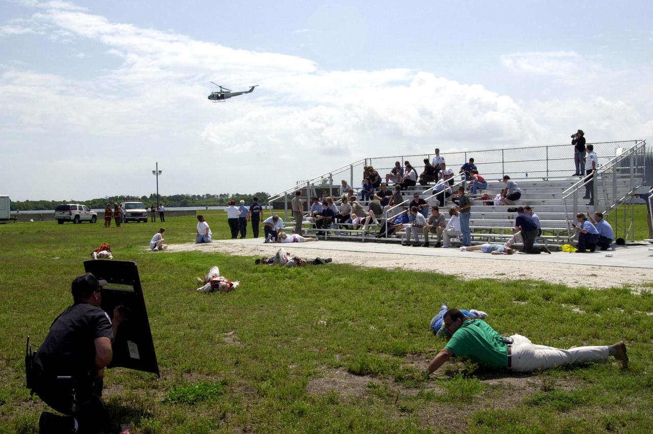 KENNEDY SPACE CENTER, FLA. -- During a staged mass casualty exercise in the Launch Complex 39 area, a security officer crouches behind a shield as if under fire by a sniper. Employees play roles of the victims (seen on the ground and in the bleachers). In the background, a helicopter arrives on the scene.The exercise was staged to validate capabilities of KSC’s fire, medical helicopter transport and security personnel to respond to such an event