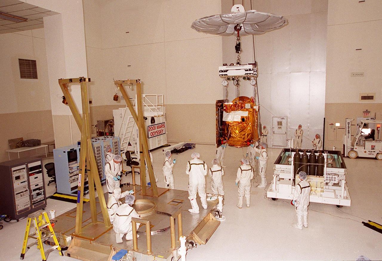 In the Spacecraft Assembly & Encapsulation Facility -2, workers help guide the <a href="http://mars.jpl.nasa.gov/2001/">2001 Mars Odyssey Orbiter </a> to a workstand (left). The spacecraft carries three science instruments: the Thermal Emission Imaging System (THEMIS), the Gamma Ray Spectrometer (GRS), and the Mars Radiation Environment Experiment (MARIE). THEMIS will map the mineralogy and morphology of the Martian surface using a high-resolution camera and a thermal infrared imaging spectrometer. The GRS will achieve global mapping of the elemental composition of the surface and determine the abundance of hydrogen in the shallow subsurface. [The GRS is a rebuild of the instrument lost with the Mars Observer mission.] The MARIE will characterize aspects of the near-space radiation environment as related to the radiation-related risk to human explorers. The Mars Odyssey Orbiter is scheduled for launch on April 7, 2001, aboard a Delta 7925 rocket from Launch Pad 17-A, Cape Canaveral Air Force Station