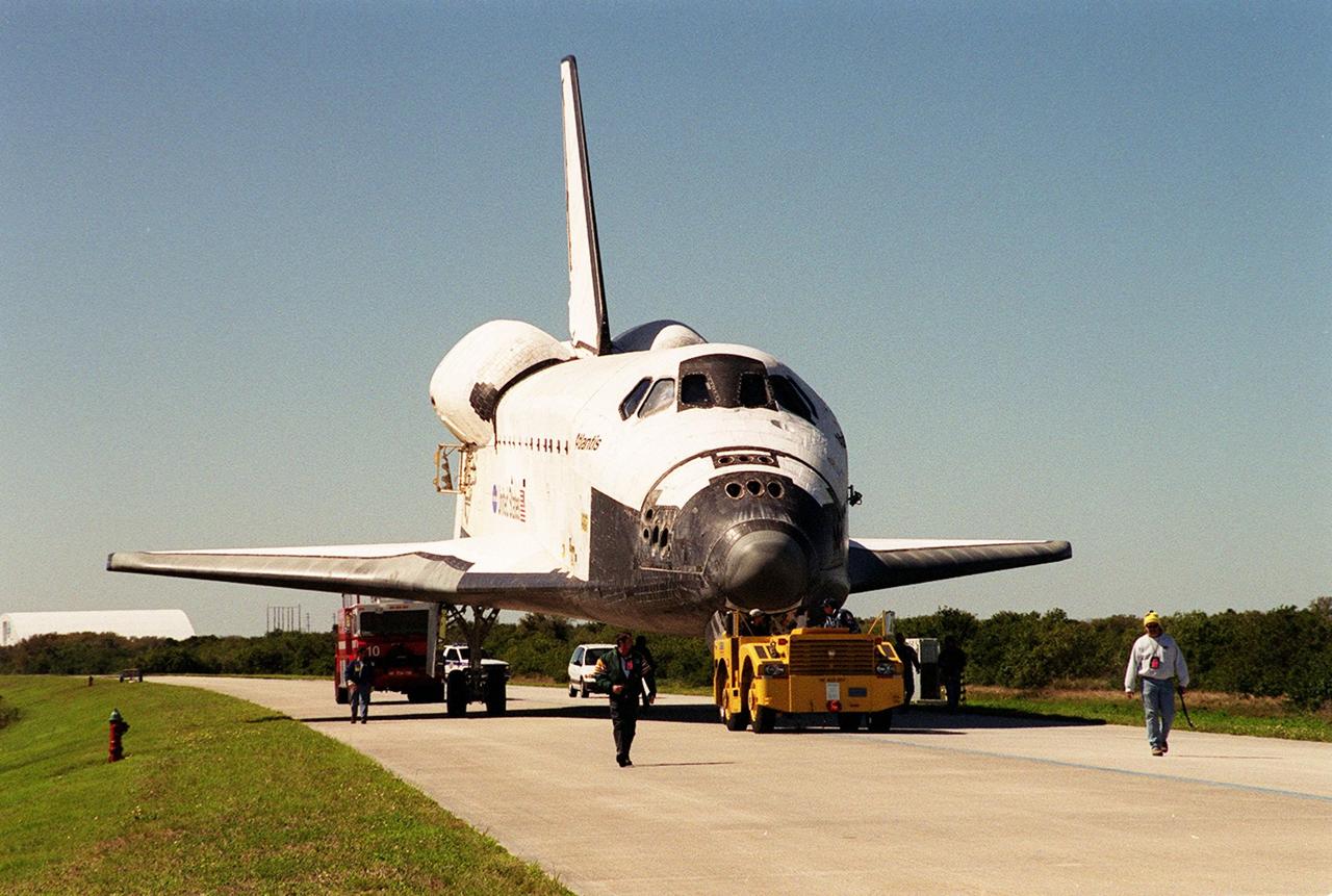 Accompanied by workers and Security, Atlantis moves along the tow-way from the KSC Shuttle Landing Facility to the Orbiter Processing Facility. Atlantis returned from California atop a Shuttle Carrier Aircraft after its Feb. 19 landing at Edwards Air Force Base, concluding mission STS-98. The ferry flight began March 1; unfavorable weather conditions kept it on the ground at Altus AFB, Okla., until it could return to Florida. In the Orbiter Processing Facility, Atlantis will be prepared for mission STS-104, the 10th construction flight to the International Space Station, scheduled to launch June 8