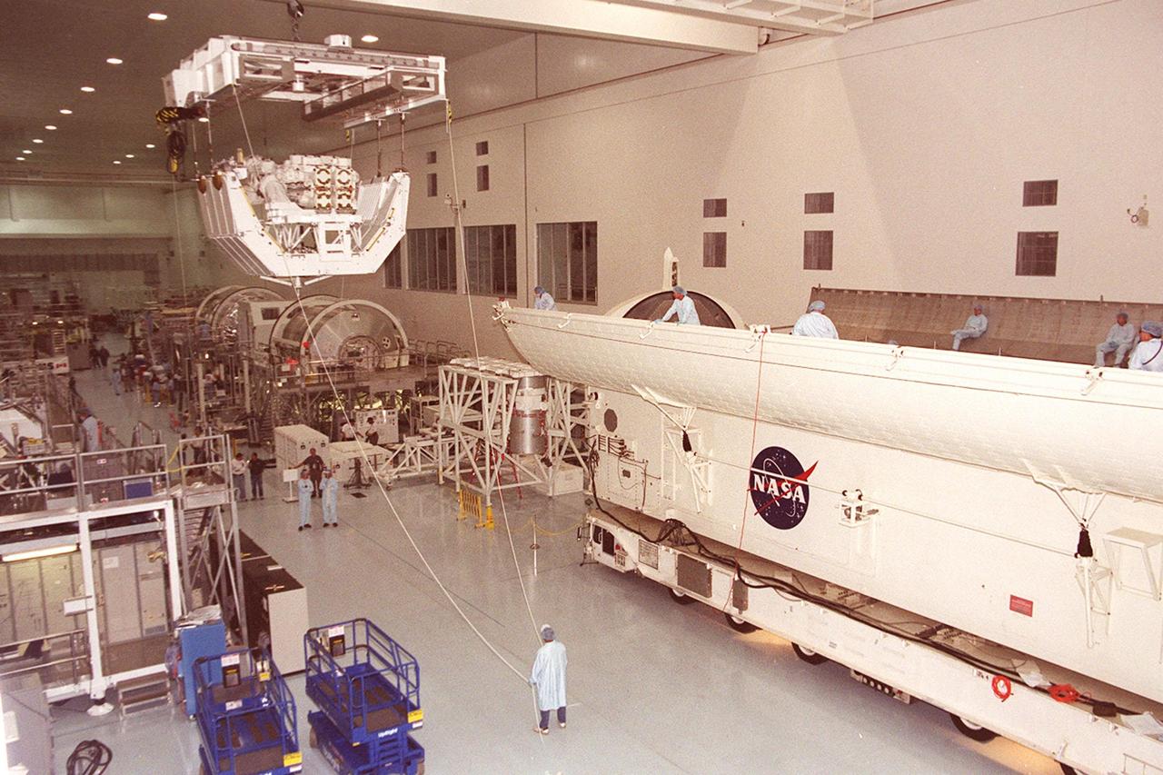 In the Space Station Processing Facility, the overhead crane carrying the pallet and Canadian robotic arm, SSRMS, nears the payload canister at right where the equipment will be placed. The arm is 57.7 feet (17.6 meters) long when fully extended and has seven motorized joints. It is capable of handling large payloads and assisting with docking the Space Shuttle. The SSRMS is self-relocatable with a Latching End Effector, so it can be attached to complementary ports spread throughout the Station’s exterior surfaces. The SSRMS is part of the payload on mission STS-100, scheduled to launch April 19 at 2:41 p.m. EDT from Launch Pad 39A, KSC