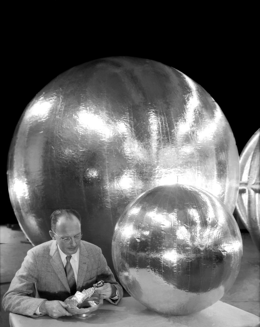 William J. O Sullivan at desk with folded subsatellite, 30 inch subsatellite, 12 foot subsatellite, and corner reflector.