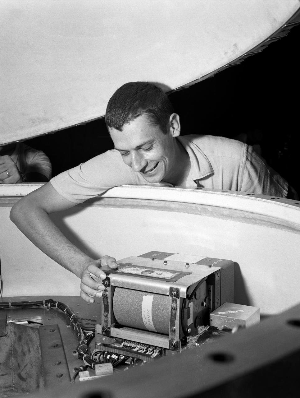 Technicians adjust the rocket motor during the attachment of the escape tower to the Mercury capsule prior to assembly with Little Joe launcher, August 20, 1959. Joseph Shortal wrote (vol. 3., p. 33):  The escape tower and rocket motors were taken from the Mercury capsule production. The tower is shown being attached to the capsule....  The escape rocket was a Grand Central 1-KS-52000 motor with three canted nozzles. The tower-jettison motor was an Atlantic Research Corp. 1.4-KS-785 motor. This was the same design tested in a beach abort test...and had the offset thrust line as used in the beach abort test to insure that the capsule would get away from the booster in an emergency. The escape system weighed 1,015 pounds, including 236 pounds of ballast for stability.   The Little Joe booster was assembled at Wallops on its special launcher in a vertical attitude. It is shown in the  on the left  with the work platform in place. The launcher was located on a special concrete slab in Launching Area 1. The capsule was lowered onto the booster by crane.... After the assembly was completed, the scaffolding was disassembled and the launcher pitched over to its normal launch angle of 80 degrees.... Little Joe had a diameter of 80 inches and an overall length, including the capsule and escape tower of 48 feet. The total weight at launch was about 43,000 pounds. The overall span of the stabilizing fins was 21.3 feet.   Although in comparison with the overall Mercury Project, Little Joe was a simple undertaking, the fact that an attempt was made to condense a normal two-year project into a 6-month one with in house labor turned it into a major undertaking for Langley.  -- Published in Joseph A. Shortal, History of Wallops Station: Origins and Activities Through 1949, (Wallops Island, VA: National Aeronautics and Space Administration, Wallops Station, nd), Comment Edition. 