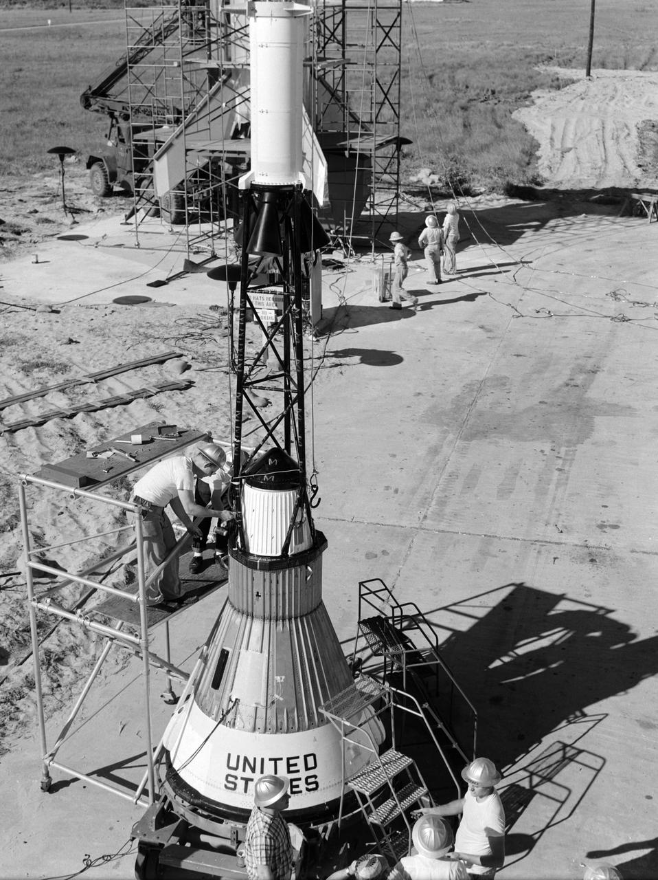 Technicians attach the escape tower to the Mercury capsule prior to assembly with Little Joe launcher, August 20, 1959. Joseph Shortal describe this as follows (vol. 3., p. 33):  The escape tower and rocket motors were taken from the Mercury capsule production. The tower is shown being attached to the capsule....  The escape rocket was a Grand Central 1-KS-52000 motor with three canted nozzles. The tower-jettison motor was an Atlantic Research Corp. 1.4-KS-785 motor. This was the same design tested in a beach abort test...and had the offset thrust line as used in the beach abort test to insure that the capsule would get away from the booster in an emergency. The escape system weighed 1,015 pounds, including 236 pounds of ballast for stability.   The Little Joe booster was assembled at Wallops on its special launcher in a vertical attitude. It is shown in the  on the left  with the work platform in place. The launcher was located on a special concrete slab in Launching Area 1. The capsule was lowered onto the booster by crane.... After the assembly was completed, the scaffolding was disassembled and the launcher pitched over to its normal launch angle of 80 degrees.... Little Joe had a diameter of 80 inches and an overall length, including the capsule and escape tower of 48 feet. The total weight at launch was about 43,000 pounds. The overall span of the stabilizing fins was 21.3 feet.   Although in comparison with the overall Mercury Project, Little Joe was a simple undertaking, the fact that an attempt was made to condense a normal two-year project into a 6-month one with in house labor turned it into a major undertaking for Langley.  -- Published in Joseph A. Shortal, History of Wallops Station: Origins and Activities Through 1949, (Wallops Island, VA: National Aeronautics and Space Administration, Wallops Station, nd), Comment Edition.