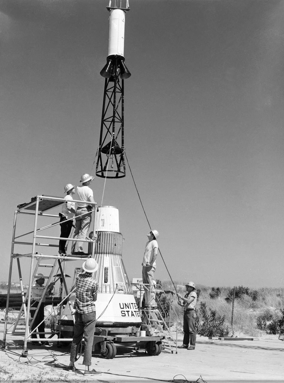  Technicians attach the escape tower to the Mercury capsule prior to assembly with Little Joe launcher, August 20, 1959. Joseph Shortal describe this as follows (vol. 3., p. 33):  The escape tower and rocket motors were taken from the Mercury capsule production. The tower is shown being attached to the capsule....  The escape rocket was a Grand Central 1-KS-52000 motor with three canted nozzles. The tower-jettison motor was an Atlantic Research Corp. 1.4-KS-785 motor. This was the same design tested in a beach abort test...and had the offset thrust line as used in the beach abort test to insure that the capsule would get away from the booster in an emergency. The escape system weighed 1,015 pounds, including 236 pounds of ballast for stability.   The Little Joe booster was assembled at Wallops on its special launcher in a vertical attitude. It is shown in the  on the left  with the work platform in place. The launcher was located on a special concrete slab in Launching Area 1. The capsule was lowered onto the booster by crane.... After the assembly was completed, the scaffolding was disassembled and the launcher pitched over to its normal launch angle of 80 degrees.... Little Joe had a diameter of 80 inches and an overall length, including the capsule and escape tower of 48 feet. The total weight at launch was about 43,000 pounds. The overall span of the stabilizing fins was 21.3 feet.   Although in comparison with the overall Mercury Project, Little Joe was a simple undertaking, the fact that an attempt was made to condense a normal two-year project into a 6-month one with in house labor turned it into a major undertaking for Langley.  -- Published in Joseph A. Shortal, History of Wallops Station: Origins and Activities Through 1949, (Wallops Island, VA: National Aeronautics and Space Administration, Wallops Station, nd), Comment Edition.