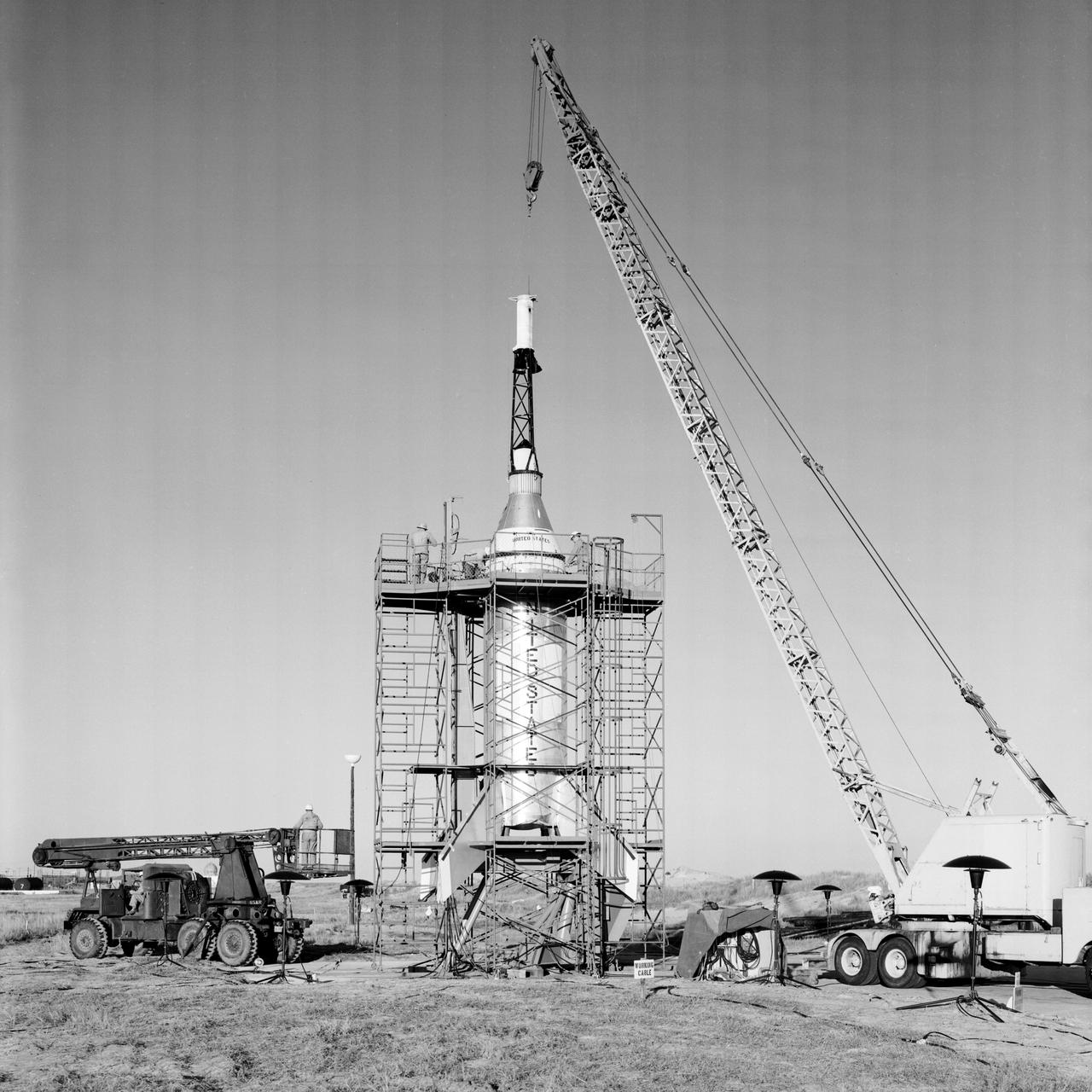 The Mercury capsule and escape tower are being lowered onto the Little Joe booster for launch on August 21, 1959. Joseph Shortal described this as follows (vol. 3, p. 33):  The Little Joe booster was assembled at Wallops on its special launcher in a vertical attitude. It is shown in the  on the left  with the work platform in place. The launcher was located on a special concrete slab in Launching Area 1. The capsule was lowered onto the booster by crane.... After the assembly was completed, the scaffolding was disassembled and the launcher pitched over to its normal launch angle of 80 degrees.... Little Joe had a diameter of 80 inches and an overall length, including the capsule and escape tower of 48 feet. The total weight at launch was about 43,000 pounds. The overall span of the stabilizing fins was 21.3 feet.   Although in comparison with the overall Mercury Project, Little Joe was a simple undertaking, the fact that an attempt was made to condense a normal two-year project into a 6-month one with in house labor turned it into a major undertaking for Langley.  -- Published in Joseph A. Shortal, History of Wallops Station: Origins and Activities Through 1949, (Wallops Island, VA: National Aeronautics and Space Administration, Wallops Station, nd), Comment Edition.