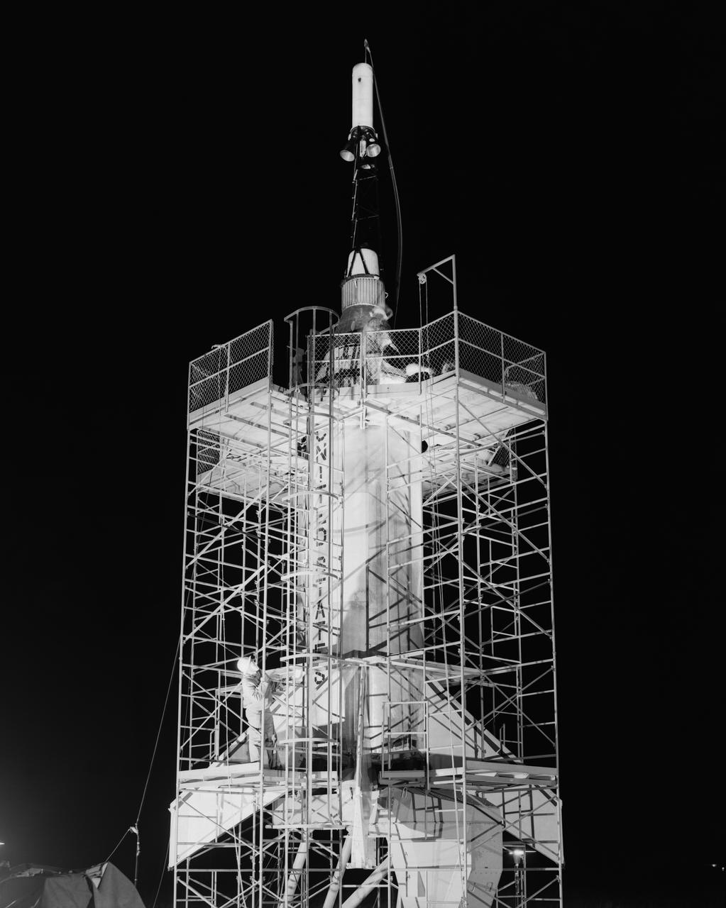 The Mercury capsule and escape tower are being lowered onto the Little Joe booster for launch on August 21, 1959. Joseph Shortal described this as follows (vol. 3, p. 33):  The Little Joe booster was assembled at Wallops on its special launcher in a vertical attitude. It is shown in the  on the left  with the work platform in place. The launcher was located on a special concrete slab in Launching Area 1. The capsule was lowered onto the booster by crane.... After the assembly was completed, the scaffolding was disassembled and the launcher pitched over to its normal launch angle of 80 degrees.... Little Joe had a diameter of 80 inches and an overall length, including the capsule and escape tower of 48 feet. The total weight at launch was about 43,000 pounds. The overall span of the stabilizing fins was 21.3 feet.   Although in comparison with the overall Mercury Project, Little Joe was a simple undertaking, the fact that an attempt was made to condense a normal two-year project into a 6-month one with in house labor turned it into a major undertaking for Langley.  -- Published in Joseph A. Shortal, History of Wallops Station: Origins and Activities Through 1949, (Wallops Island, VA: National Aeronautics and Space Administration, Wallops Station, nd), Comment Edition.