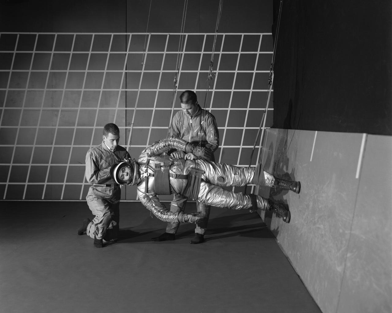 A test subject being suited up for studies on the Reduced Gravity Walking Simulator located in the hangar at Langley Research Center. The initial version of this simulator was located inside the hangar. Later a larger version would be located at the Lunar Landing Facility. The purpose of this simulator was to study the subject while walking, jumping or running. Researchers conducted studies of various factors such as fatigue limit, energy expenditure, and speed of locomotion. Francis B. Smith wrote in his paper "Simulators For Manned Space Research," "I would like to conclude this talk with a discussion of a device for simulating lunar gravity which is very effective and yet which is so simple that its cost is in the order of a few thousand dollars at most, rather than hundreds of thousands. With a little ingenuity, one could almost build this type simulator in his backyard for children to play on. The principle is ...if a test subject is suspended in a sling so that his body axis makes an angle of 9 1/2 degrees with the horizontal and if he then "stands" on a platform perpendicular to his body axis, the component of the earth's gravity forcing him toward the platform is one times the sine of 9 1/2 degrees or approximately 1/6 of the earth's normal gravity field. That is, a 180 pound astronaut "standing" on the platform would exert a force of only 30 pounds - the same as if he were standing upright on the lunar surface." -- Published in James R. Hansen, Spaceflight Revolution: NASA Langley Research Center From Sputnik to Apollo, NASA SP-4308; Francis B. Smith, "Simulators For Manned Space Research," Paper for 1966 IEEE International Convention, New York, NY, March 21-25, 1966