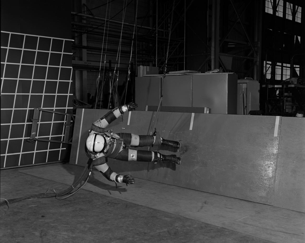 A "suited" test subject on the Reduced Gravity Walking Simulator located in the hangar at Langley Research Center. The initial version of this simulator was located inside the hangar. Later a larger version would be located at the Lunar Landing Facility. The purpose of this simulator was to study the subject while walking, jumping or running. Researchers conducted studies of various factors such as fatigue limit, energy expenditure, and speed of locomotion. Francis B. Smith wrote in "Simulators For Manned Space Research:" "The cables which support the astronaut are supported by an overhead trolley about 150 feet above the center line of the walkway and the support is arranged so that the subject is free to walk, run, jump, and perform other self-locomotive tasks in a more-or-less normal manner, even though he is constrained to move in one place." "The studies thus far show that an astronaut should have no particular difficulty in walking in a pressurized space suit on a hard lunar surface. Rather, the pace was faster and the suit was found to be more comfortable and less fatiguing under lunar "g" than under earth "g." When the test subject wished to travel hurriedly any appreciable distance, a long loping gait at about 10 feet per second was found to be most comfortable." -- Published in James R. Hansen, Spaceflight Revolution: NASA Langley Research Center From Sputnik to Apollo, (Washington: NASA, 1995), p. 377; Francis B. Smith, "Simulators For Manned Space Research," Paper for 1966 IEEE International Convention, New York, NY, March 21-25, 1966.