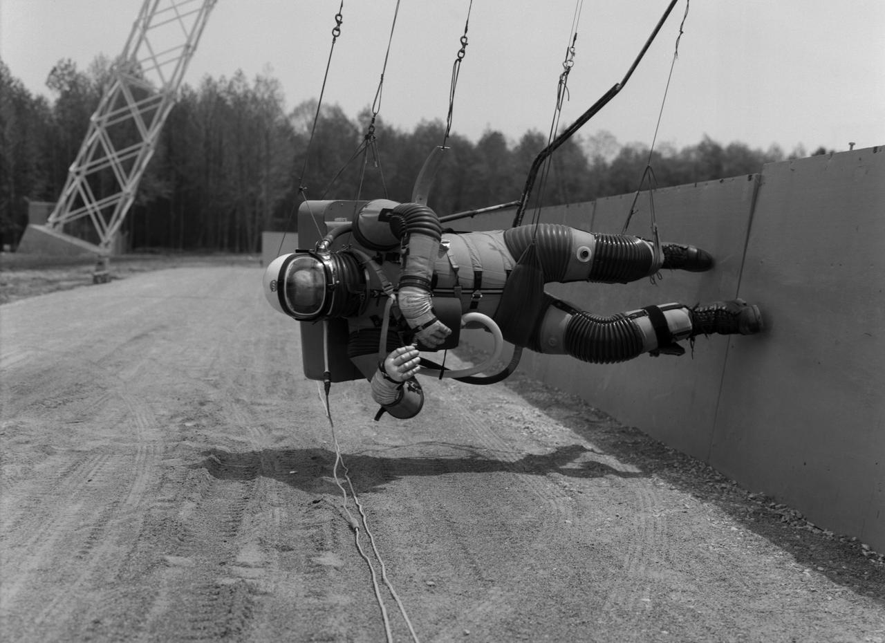 Test subject wearing the pressurized "space" suit for the Reduced Gravity Walking Simulator located at the Lunar Landing Facility. The purpose of this simulator was to study the subject while walking, jumping or running. Researchers conducted studies of various factors such as fatigue limit, energy expenditure, and speed of locomotion. A.W. Vigil described the purpose of the simulator in his paper "Discussion of Existing and Planned Simulators for Space Research," "When the astronauts land on the moon they will be in an unfamiliar environment involving, particularly, a gravitational field only one-sixth as strong as on earth. A novel method of simulating lunar gravity has been developed and is supported by a puppet-type suspension system at the end of a long pendulum. A floor is provided at the proper angle so that one-sixth of the subject's weight is supported by the floor with the remainder being supported by the suspension system. This simulator allows almost complete freedom in vertical translation and pitch and is considered to be a very realistic simulation of the lunar walking problem. For this problem this simulator suffers only slightly from the restrictions in lateral movement it puts on the test subject. This is not considered a strong disadvantage for ordinary walking problems since most of the motions do, in fact, occur in the vertical plane. However, this simulation technique would be severely restrictive if applied to the study of the extra-vehicular locomotion problem, for example, because in this situation complete six degrees of freedom are rather necessary. This technique, in effect, automatically introduces a two-axis attitude stabilization system into the problem. The technique could, however, be used in preliminary studies of extra-vehicular locomotion where, for example, it might be assumed that one axis of the attitude control system on the astronaut maneuvering unit may have failed." -- Published in James R. Hansen, Spaceflight Revolution: NASA Langley Research Center From Sputnik to Apollo, (Washington: NASA, 1995), p. 377; A.W. Vigil, "Discussion of Existing and Planned Simulators for Space Research," Paper presented at Conference on the Role of Simulation in Space Technology," Blacksburg, VA, August 17-21, 1964.