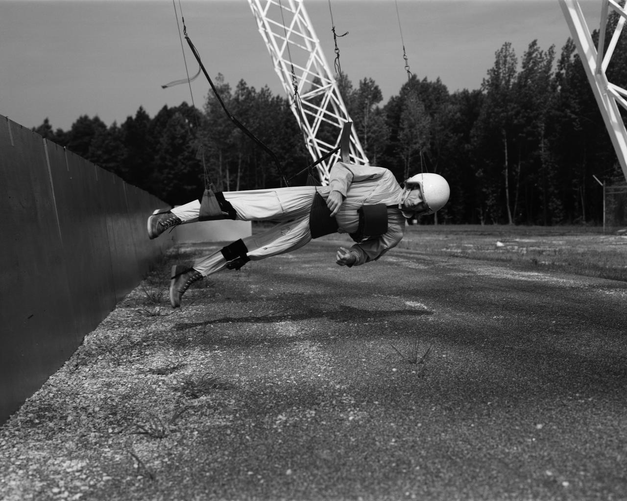 Astronaut Walt Cunningham on the Reduced Gravity Walking Simulator located at the Lunar Landing Facility. The purpose of this simulator was to study the subject while walking, jumping or running. Researchers conducted studies of various factors such as fatigue limit, energy expenditure, and speed of locomotion. A.W. Vigil described the purpose of the simulator in his paper "Discussion of Existing and Planned Simulators for Space Research," "When the astronauts land on the moon they will be in an unfamiliar environment involving, particularly, a gravitational field only one-sixth as strong as on earth. A novel method of simulating lunar gravity has been developed and is supported by a puppet-type suspension system at the end of a long pendulum. A floor is provided at the proper angle so that one-sixth of the subject's weight is supported by the floor with the remainder being supported by the suspension system. This simulator allows almost complete freedom in vertical translation and pitch and is considered to be a very realistic simulation of the lunar walking problem. For this problem this simulator suffers only slightly from the restrictions in lateral movement it puts on the test subject. This is not considered a strong disadvantage for ordinary walking problems since most of the motions do, in fact, occur in the vertical plane. However, this simulation technique would be severely restrictive if applied to the study of the extra-vehicular locomotion problem, for example, because in this situation complete six degrees of freedom are rather necessary. This technique, in effect, automatically introduces a two-axis attitude stabilization system into the problem. The technique could, however, be used in preliminary studies of extra-vehicular locomotion where, for example, it might be assumed that one axis of the attitude control system on the astronaut maneuvering unit may have failed." -- Published in James R. Hansen, Spaceflight Revolution: NASA Langley Research Center From Sputnik to Apollo, (Washington: NASA, 1995), p. 377; A.W. Vigil, "Discussion of Existing and Planned Simulators for Space Research," Paper presented at Conference on the Role of Simulation in Space Technology," Blacksburg, VA, August 17-21, 1964.