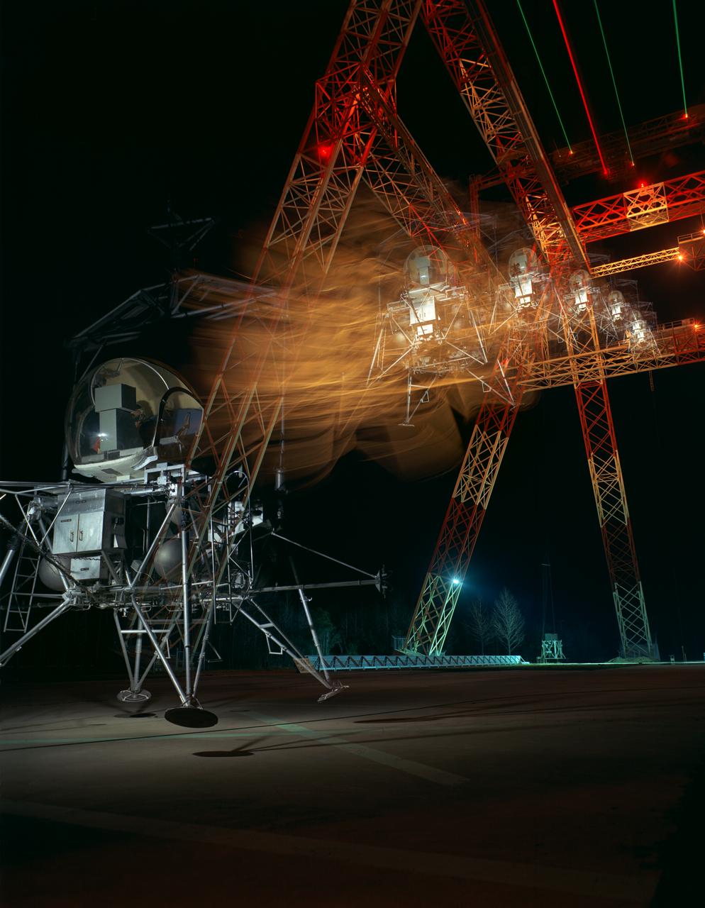 During a nighttime training session, a multiple exposure captures the movement of the Lunar Excursion Module Simulator (LEMS). The LEMS was a manned vehicle used to familiarize the Apollo astronauts with the handling characteristics of lunar-landing type vehicle.  The Apollo Program is best known for the astronaut Neal Armstrong s first step on the Moon July 20, 1969. In its earliest test period, the LEMS featured a helicopter crew cabin atop the lunar landing module. Later, the helicopter crew cabin was replaced with a stand-up rectangular cabin which was more efficient for controlling maneuvers and for better viewing by the pilot.  The vehicle was designed at Langley Research Center in Hampton, VA.  This multiple exposure shows a simulated Moon landing of the (LEMS) trainer at Langley s Lunar Landing Research Facility.  -- Photograph published in Winds of Change, 75th Anniversary NASA publication (page 70), by James Shultz. Also published in  " A Century at Langley" by Joseph Chambers, pg. 93. 