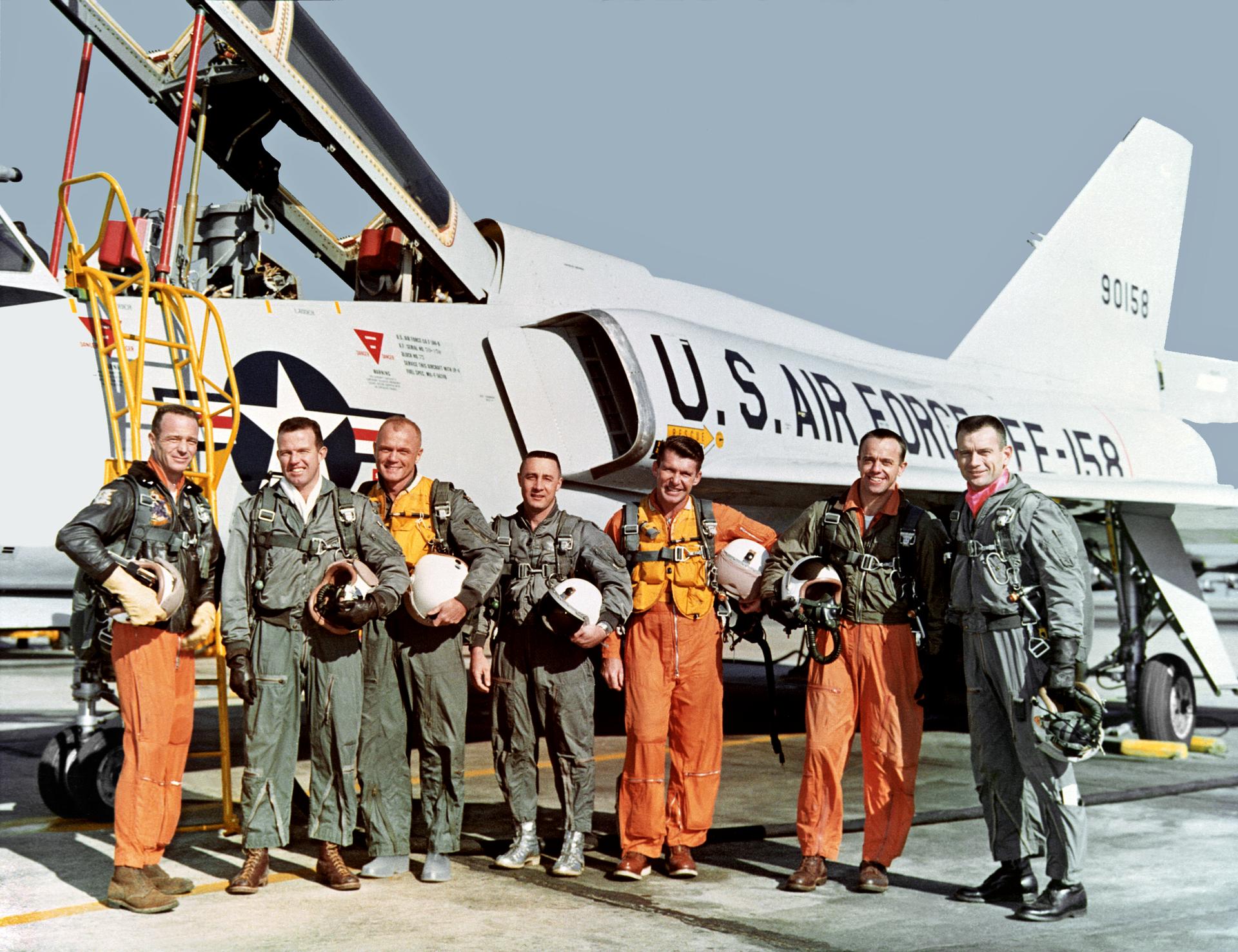 The original seven astronauts standing in front of a jet airplane all wearing flight suits. The airplane has the Airforce insignia on the side with the cockpit open.