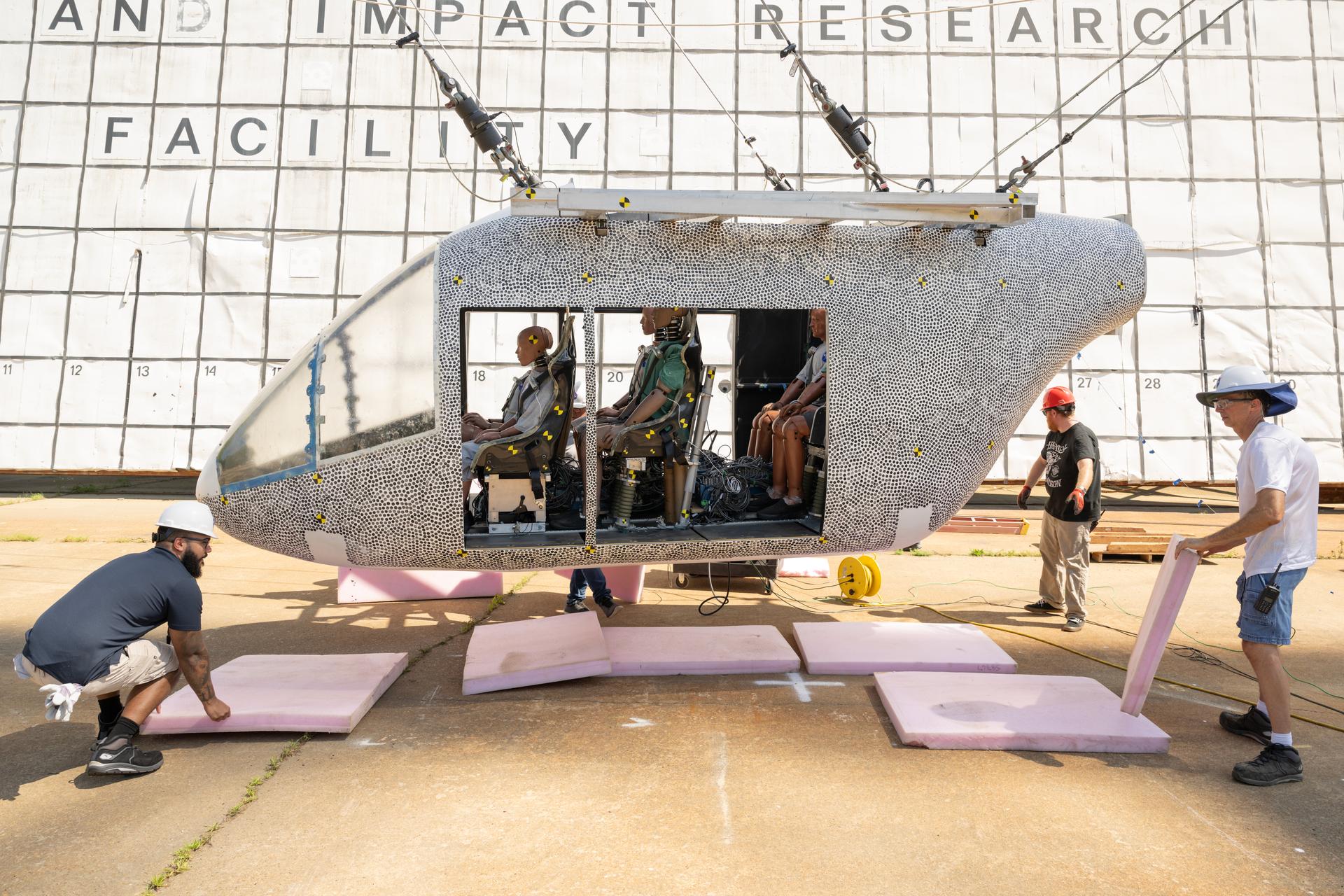 A white aircraft body with small black dots is hoisted in the air by cables. There are several test dummies inside of the aircraft. The aircraft does not hang far off of the ground. Three workers with hardhats remove pink foam squares from underneath the aircraft. Behind the aircraft is a large white board to measure the height, which includes many black squares with small numbers inside some of the bottom squares.