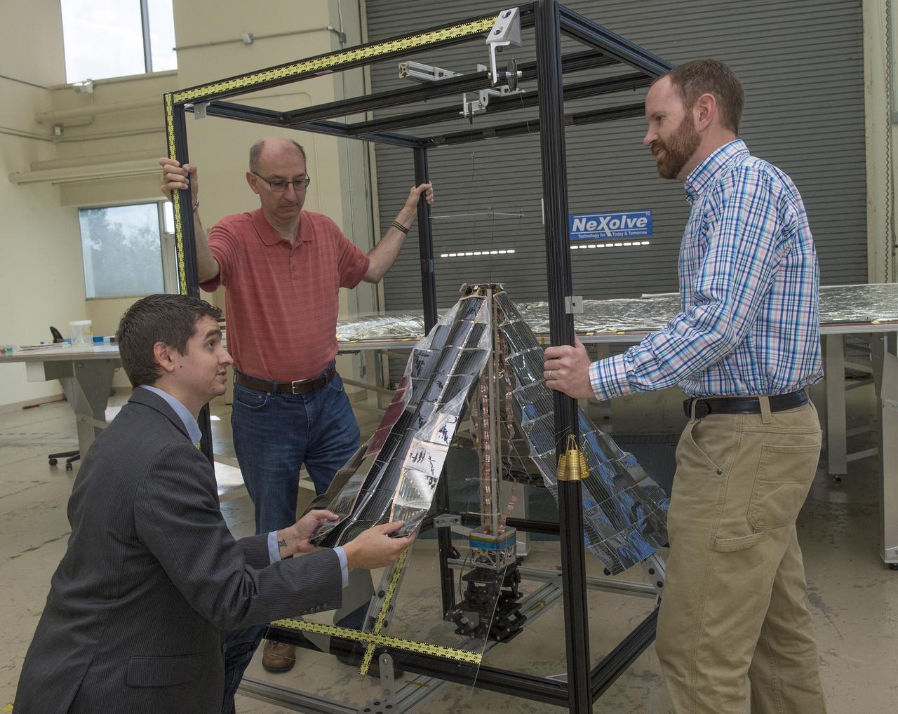 JOHN CARR, CO-PRINCIPAL INVESTIGATOR FOR NASA'S LIGHTWEIGHT INTEGRATED SOLAR ARRAY AND TRANSCEIVER PROJECT, KNEELS TO SHOW HOW ONE OF THE THIN-FILM SIDES OR "PETALS" IN WHICH PHOTO-VOLTAIC CELLS ARE EMBEDDED, IS FOLDED AND STOWED BEFORE LAUNCH. LOOKING ON DURING A DEMONSTRATION AFTER TESTING AT NEXOLVE, ARE LES JOHNSON, LEFT, ALSO CO-PRINCIPAL INVESTIGATOR, AND DARREN BOYD, RIGHT, THE RADIO FREQUENCY LEAD FOR THE PROJECT.