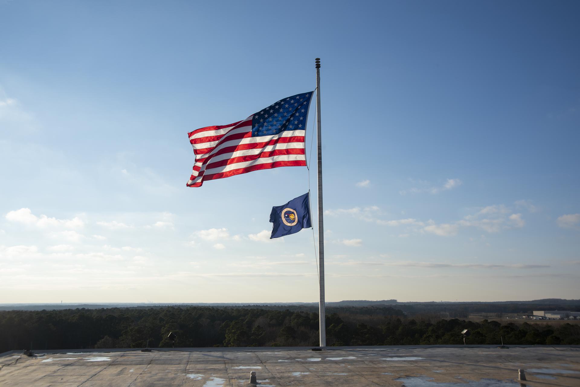 A US Flag and NASA flag are waving in the wind against a bright blue sky.