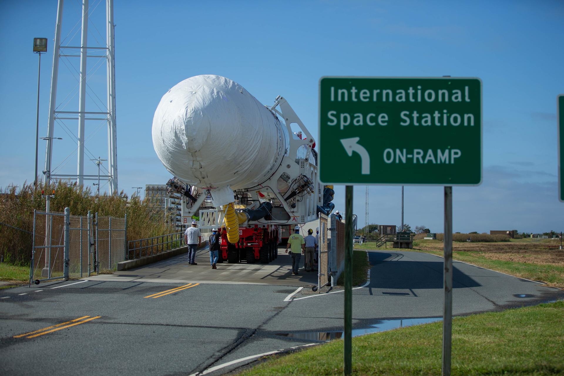 A white Northrop Grumman Antares rocket is rolled horizontally to the launch pad. A green and white road sign in front of the rocket reads "International Space Station On-ramp" with an arrow pointing to the launch pad.