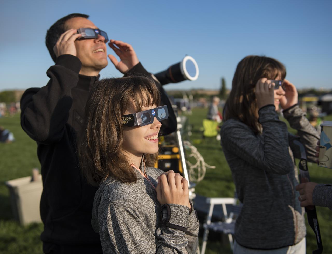 Klaus Koschinsky, left, and his daughters, Claudia, center, and Tanja, right, from Munich, Germany, are seen as they watch a total solar eclipse through protective glasses in Madras, Oregon on Monday, Aug. 21, 2017. A total solar eclipse swept across a narrow portion of the contiguous United States from Lincoln Beach, Oregon to Charleston, South Carolina. A partial solar eclipse was visible across the entire North American continent along with parts of South America, Africa, and Europe.  Photo Credit: (NASA/Aubrey Gemignani)