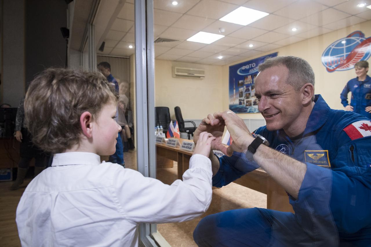Expedition 58 Flight Engineer David Saint-Jacques of the Canadian Space Agency (CSA), right, who is in quarantine, makes a heart with his hands for his son, at the conclusion of a press conference, Sunday, Dec. 2, 2018 at the Cosmonaut Hotel in Baikonur, Kazakhstan. Launch of the Soyuz rocket is scheduled for Dec. 3 and will carry Saint-Jacques, Soyuz Commander Oleg Kononenko of Roscosmos, and Flight Engineer Anne McClain of NASA into orbit to begin their six and a half month mission on the International Space Station. Photo Credit: (NASA/Aubrey Gemignani).