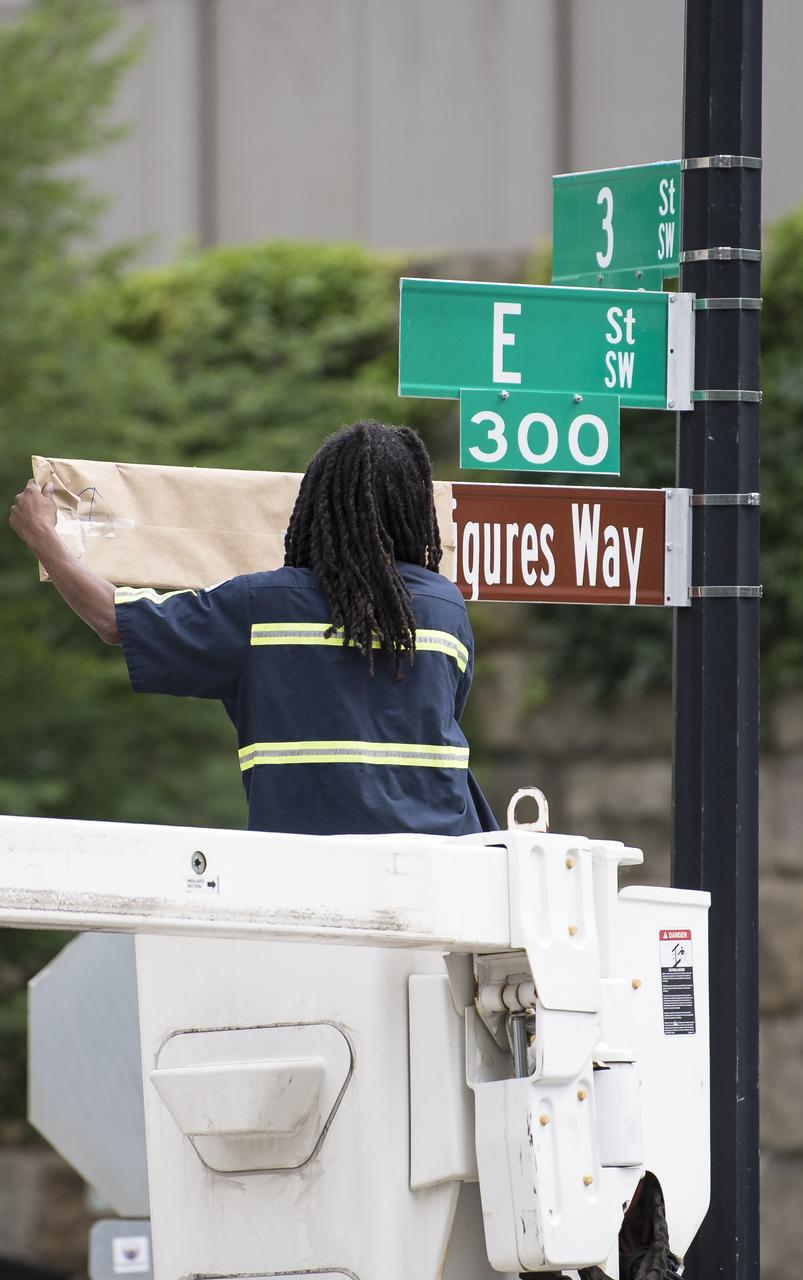 A D.C. Department of Transportation employee removes a paper cover from the "Hidden Figures Way" street sign at the corner of 3rd and E Street SW during a dedication ceremony, Wednesday, June 12, 2019 at NASA Headquarters in Washington, DC. The 300 block of E Street SW in front of the NASA Headquarters building was designated as "Hidden Figures Way" to honor Katherine Johnson, Dorothy Vaughan, Mary Jackson and all women who have dedicated their lives to honorably serving their country, advancing equality, and contributing to the space program of the United States. Photo Credit: (NASA/Joel Kowsky)