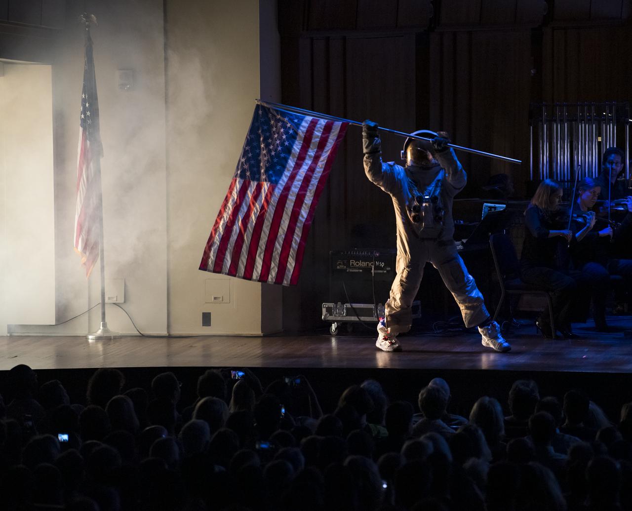 Maker and host of Savage Builds, Adam Savage, dressed in a spacesuit, plants a U.S. flag on the stage during the "National Symphony Orchestra Pops, Apollo 11: A 50th Anniversary, One Small Step, One Giant Leap" a program including musical acts, speakers, and images and video related to space, on Saturday, July 20, 2019 at the John F. Kennedy Center for the Performing Arts in Washington. NASA and the country are recognizing the 50th anniversary of Apollo 11, in which astronauts Neil Armstrong, Michael Collins, and Buzz Aldrin crewed the first mission to land astronauts on the Moon. Photo Credit: (NASA/Aubrey Gemignani)