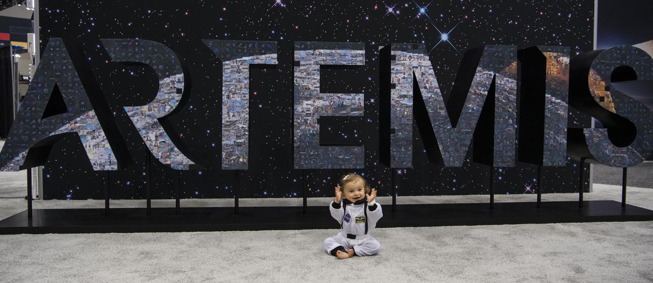 A baby in a flight suit sits in front of the Artemis sign at the NASA exhibit during the 70th International Astronautical Congress, Friday, Oct. 25, 2019, at the Walter E. Washington Convention Center in Washington. Photo credit: (NASA/Aubrey Gemignani)