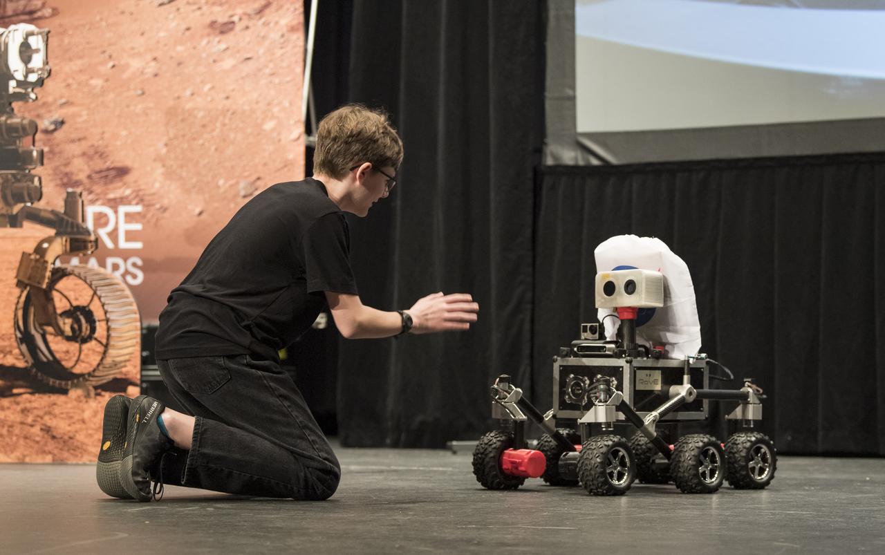 ROV-E, a new Mars outreach rover, rolls toward Alex Mather, the student whose submission, Perseverance, was chosen as the official name of the Mars 2020 rover, with NASA handouts at an event to announce the official name of the rover, Thursday, March 5, 2020, at Lake Braddock Secondary School in Burke, Va. The final selection of the new name, Perseverance, was made by Associate Administrator of NASA's Science Mission Directorate, Thomas Zurbuchen, following a nationwide naming contest conducted in 2019 that drew more than 28,000 essays by K-12 students from every U.S. state and territory. The rover is currently at NASA's Kennedy Space Center in Florida being prepared for launch this summer. Photo Credit: (NASA/Aubrey Gemignani)