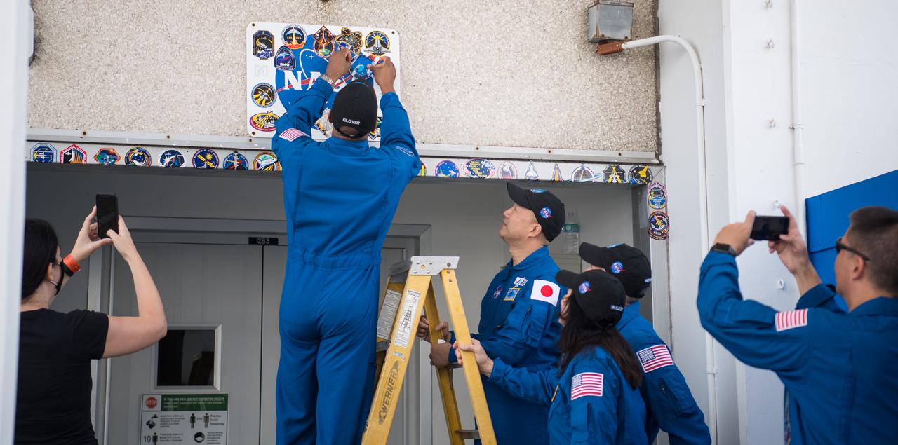 NASA astronaut Victor Glover, left on ladder, places a Crew-1 mission sticker above the doorway to crew quarters at the Neil  A. Armstrong Operations and Checkout Building as fellow crewmates astronauts Soichi Noguchi of the Japan Aerospace Exploration Agency (JAXA), and Shannon Walker and Mike Hopkins of NASA look on, Thursday, Nov. 12, 2020, at NASA’s Kennedy Space Center in Florida. NASA’s SpaceX Crew-1 mission is the first crew rotation mission of the SpaceX Crew Dragon spacecraft and Falcon 9 rocket to the International Space Station as part of the agency’s Commercial Crew Program. Glover, Noguchi, Walker, and Hopkins are scheduled to launch at 7:27 p.m. EST on Sunday, Nov. 15, from Launch Complex 39A at the Kennedy Space Center. Photo Credit: (NASA/Joel Kowsky)