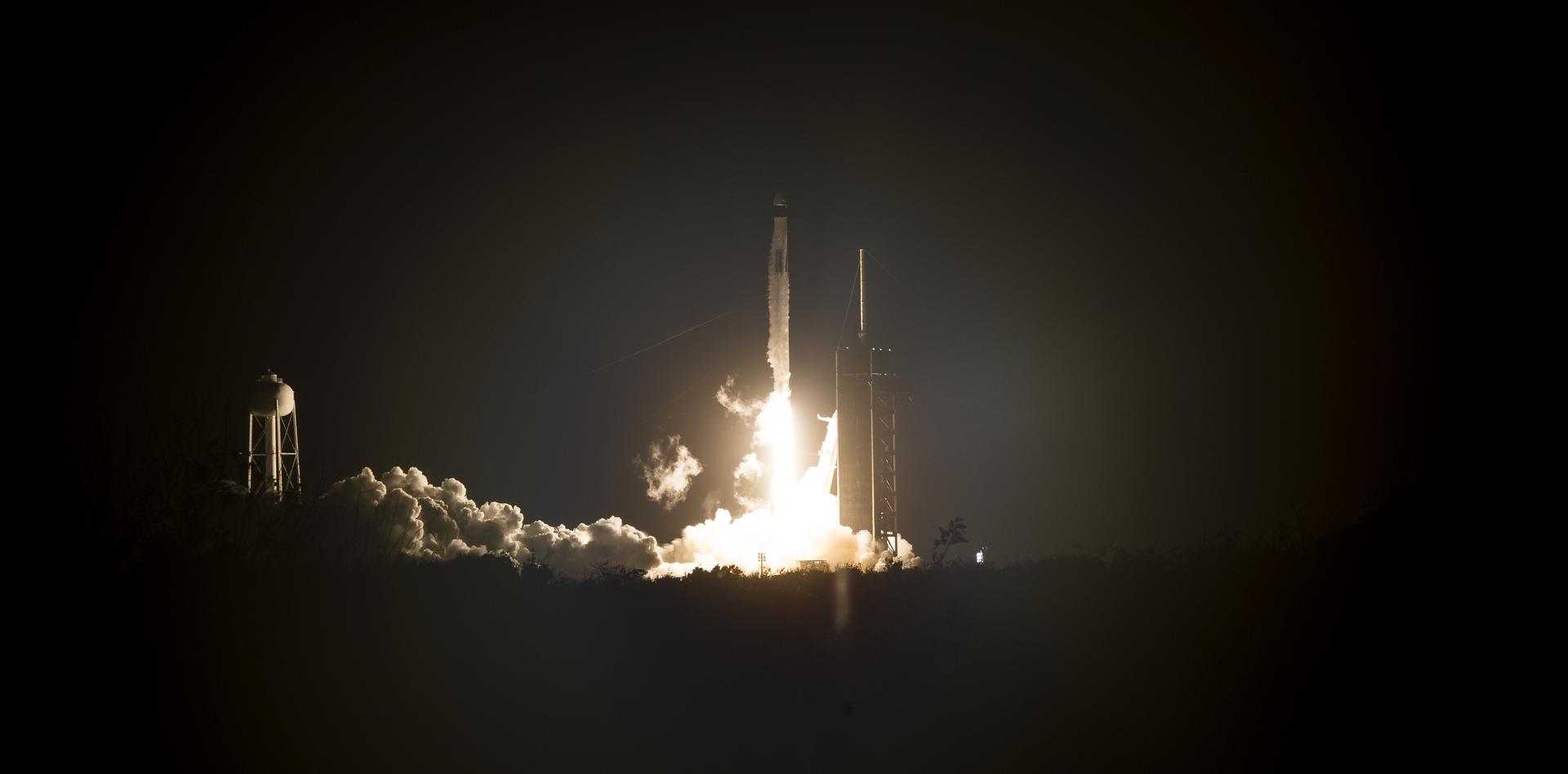 In the center of the image, a rocket lifts into a dark night sky above a column of bright fire and smoke billows out to the left. The launch tower is visible to the right of the fire column.