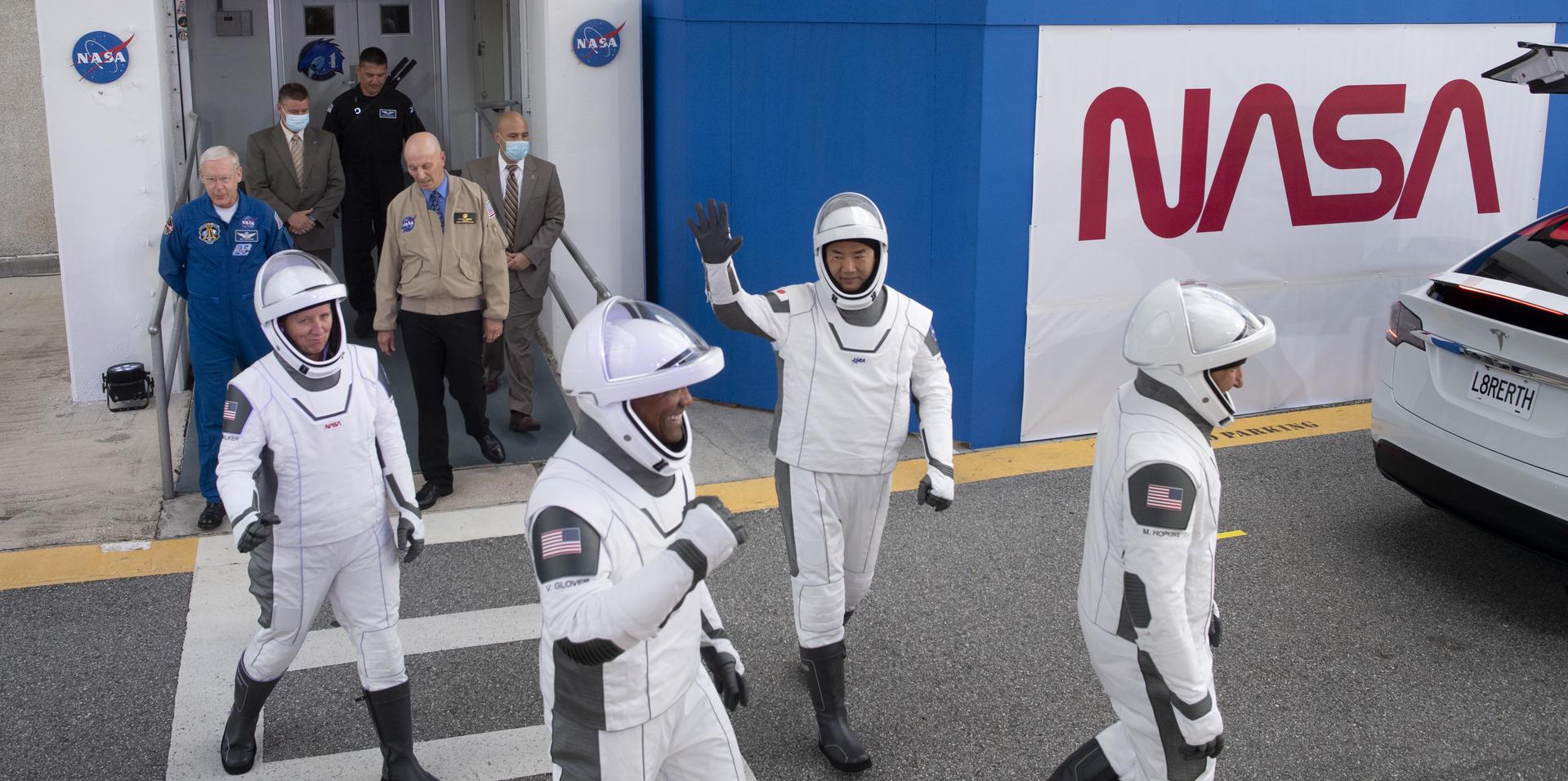 Four astronauts in their spacesuits wave to crowd with NASA insignia in the background