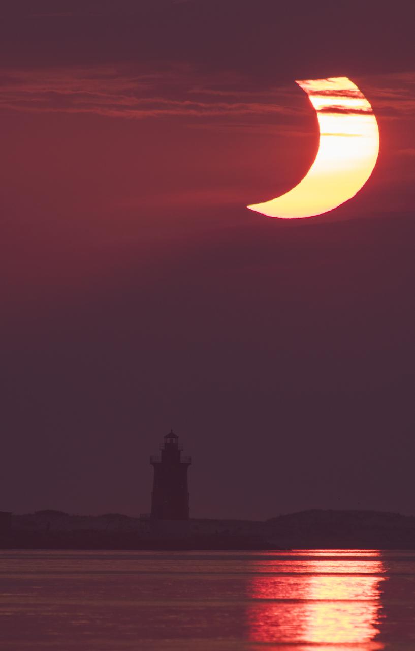 A partial solar eclipse is seen as the sun rises behind the Delaware Breakwater Lighthouse, Thursday, June 10, 2021, at Lewes Beach in Delaware. The annular or “ring of fire” solar eclipse is only visible to some parts of Greenland, Northern Russia, and Canada. Photo Credit: (NASA/Aubrey Gemignani)