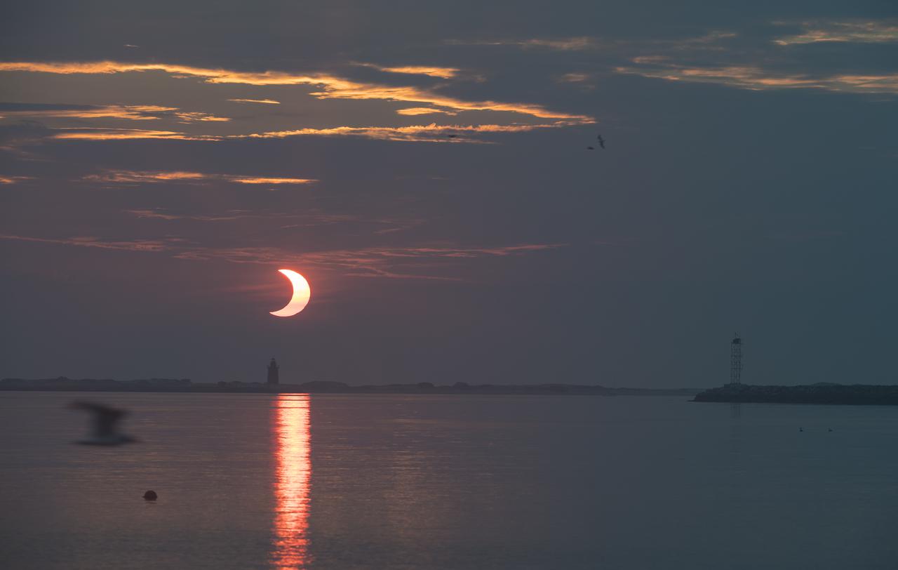 A partial solar eclipse is seen as the sun rises behind the Delaware Breakwater Lighthouse, Thursday, June 10, 2021, at Lewes Beach in Delaware. The annular or “ring of fire” solar eclipse is only visible to some parts of Greenland, Northern Russia, and Canada. Photo Credit: (NASA/Aubrey Gemignani)