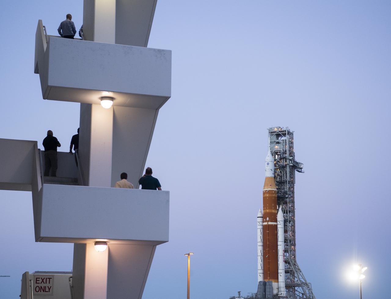 NASA employees are seen on the stairs outside of the Rocco A. Petrone Launch Control Center as NASA’s Space Launch System (SLS) rocket with the Orion spacecraft aboard atop a mobile launcher rolls out of High Bay 3 of the Vehicle Assembly Building for the first time to Launch Complex 39B, Thursday, March 17, 2022, at NASA’s Kennedy Space Center in Florida. Ahead of NASA’s Artemis I flight test, the fully stacked and integrated SLS rocket and Orion spacecraft will undergo a wet dress rehearsal at Launch Complex 39B to verify systems and practice countdown procedures for the first launch. Photo Credit: (NASA/Joel Kowsky)