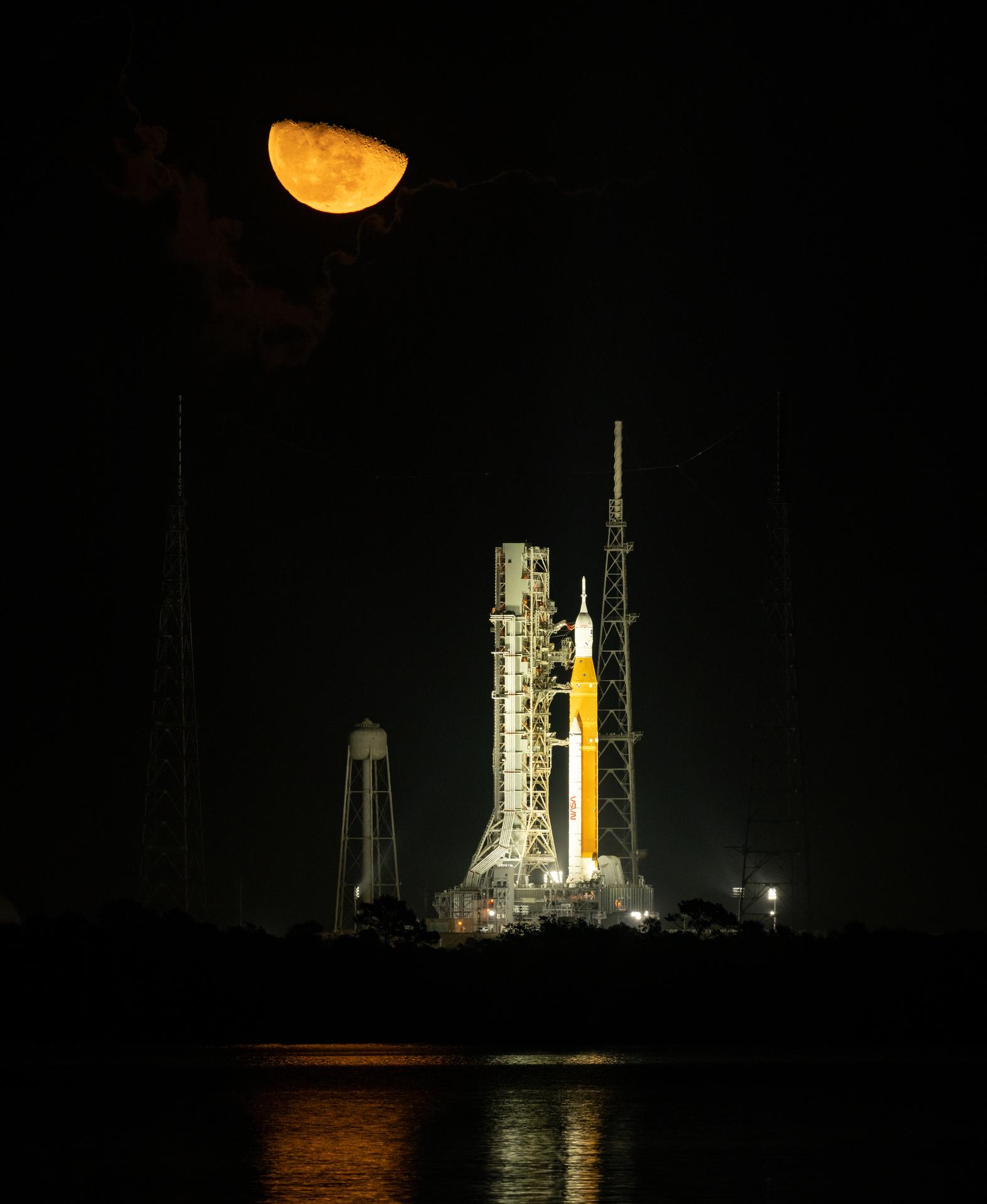 Amidst a dark nighttime sky, a golden-hued moon is seen rising above NASA’s Space Launch System (SLS) rocket with the Orion spacecraft aboard at Launch Pad 39B at NASA’s Kennedy Space Center in Florida.