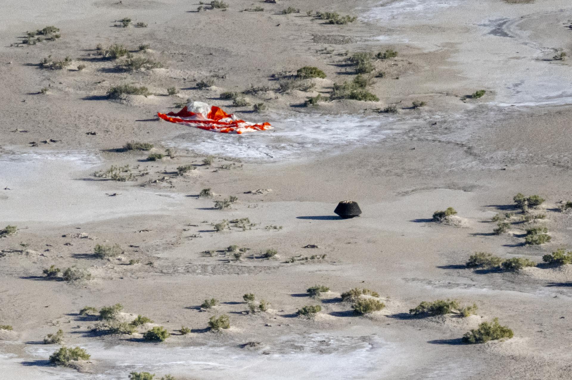 Aerial view of the OSIRIS-REx sample return capsule on the desert ground. An orange-and-white parachute lays crumpled next to it.⁣