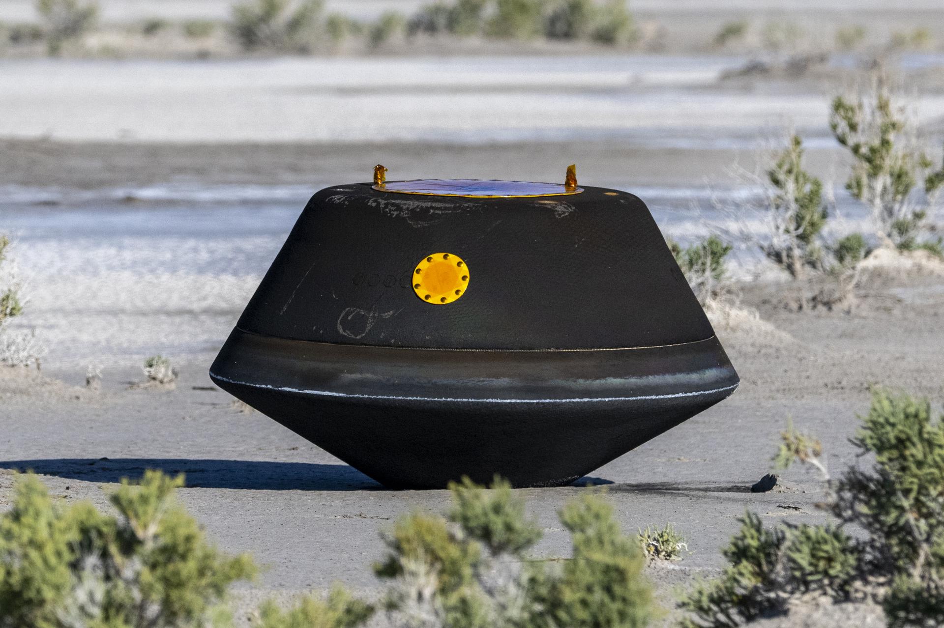 Black, top-shaped sample return capsule sits on the desert ground.