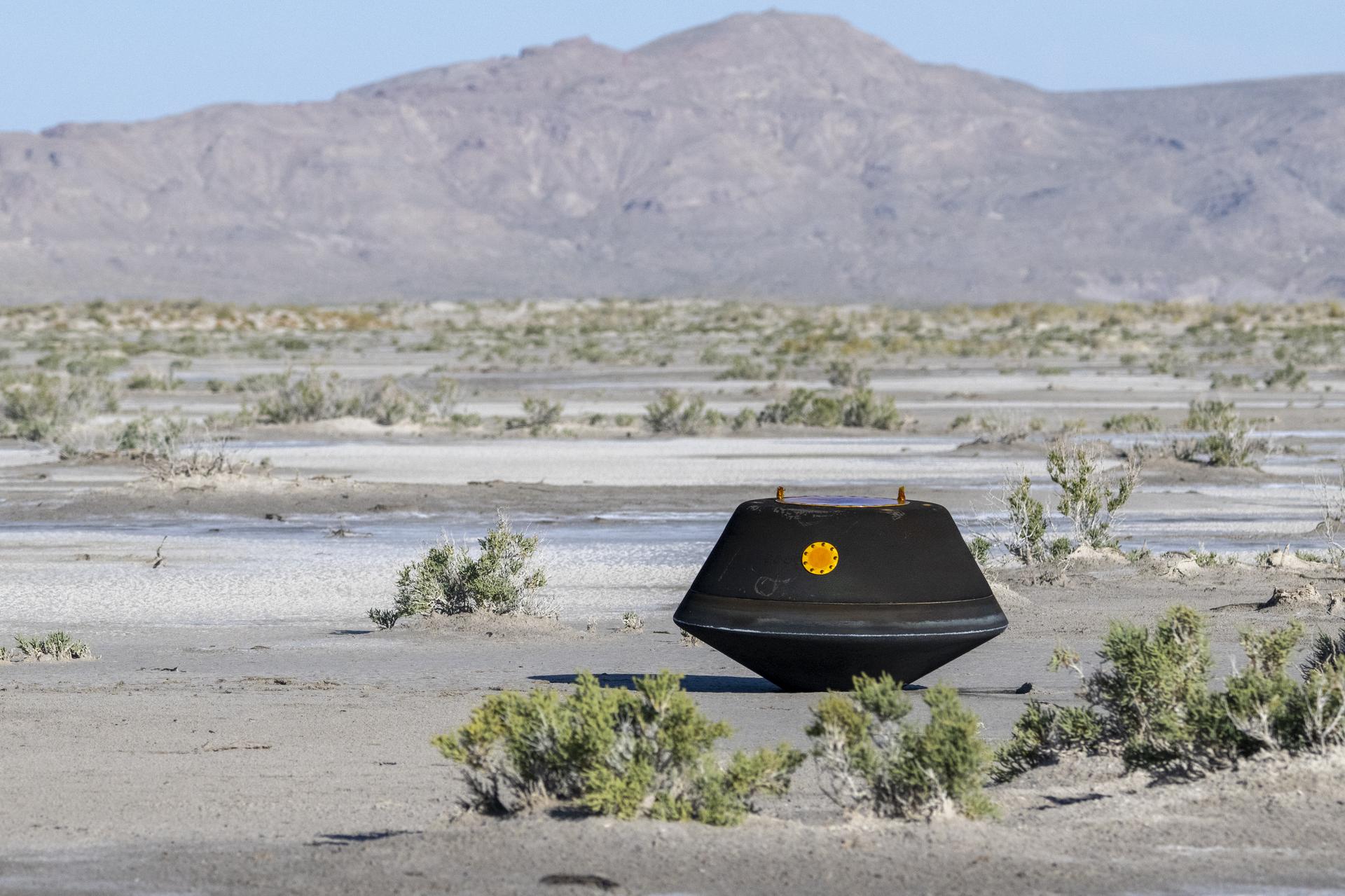 Black, top-shaped sample return capsule sits on the desert ground.