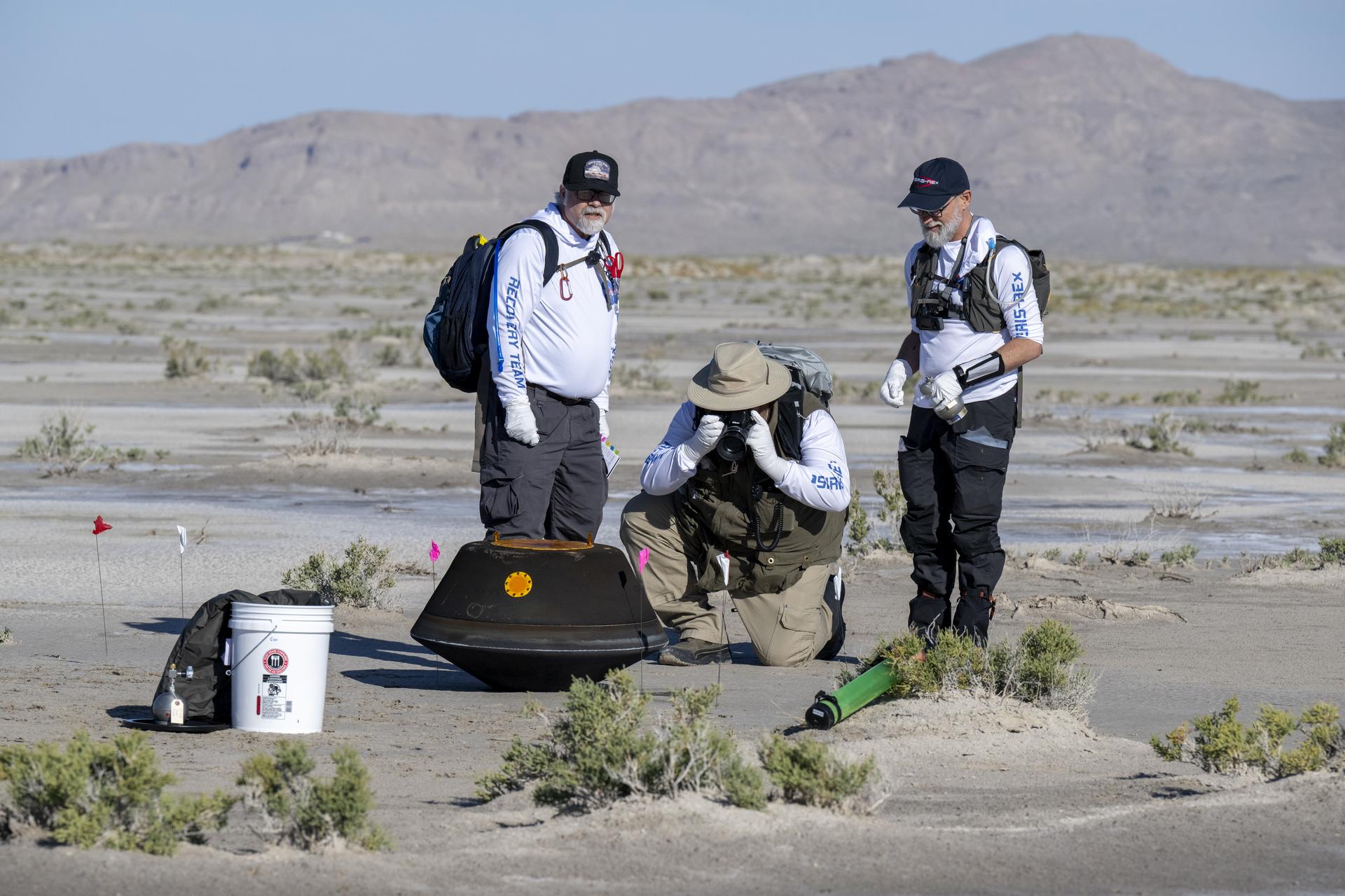 Three men look at the OSIRIS-REx sample capsule in the desert. One is kneeling, two are standing. 